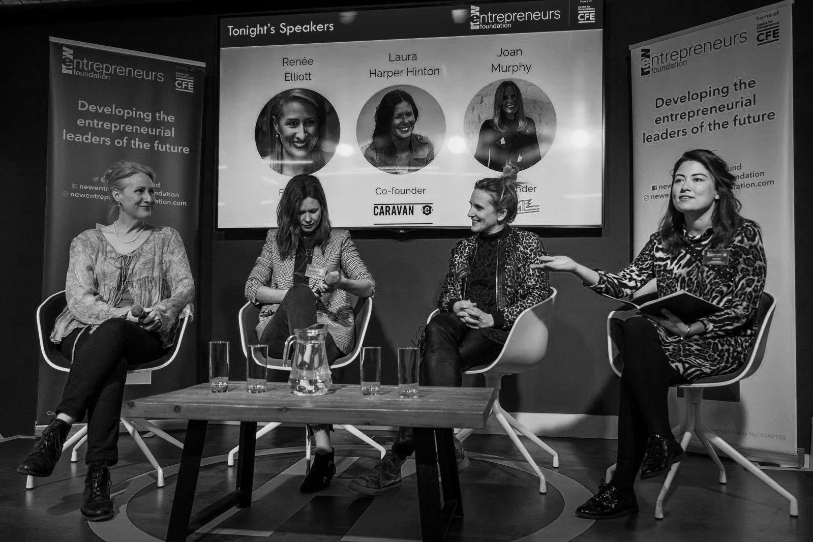 Four women participating in a panel discussion at an event on entrepreneurship, with a screen displaying their names and photos behind them. Banners on each side display the event's theme about developing future entrepreneurial leaders.