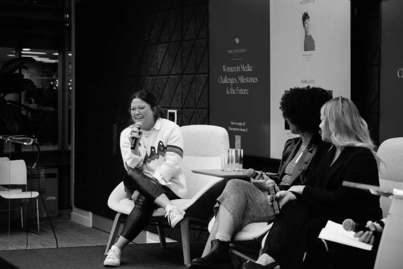 A woman speaking into a microphone on stage at a panel discussion with two other women, in a room with posters on the wall.