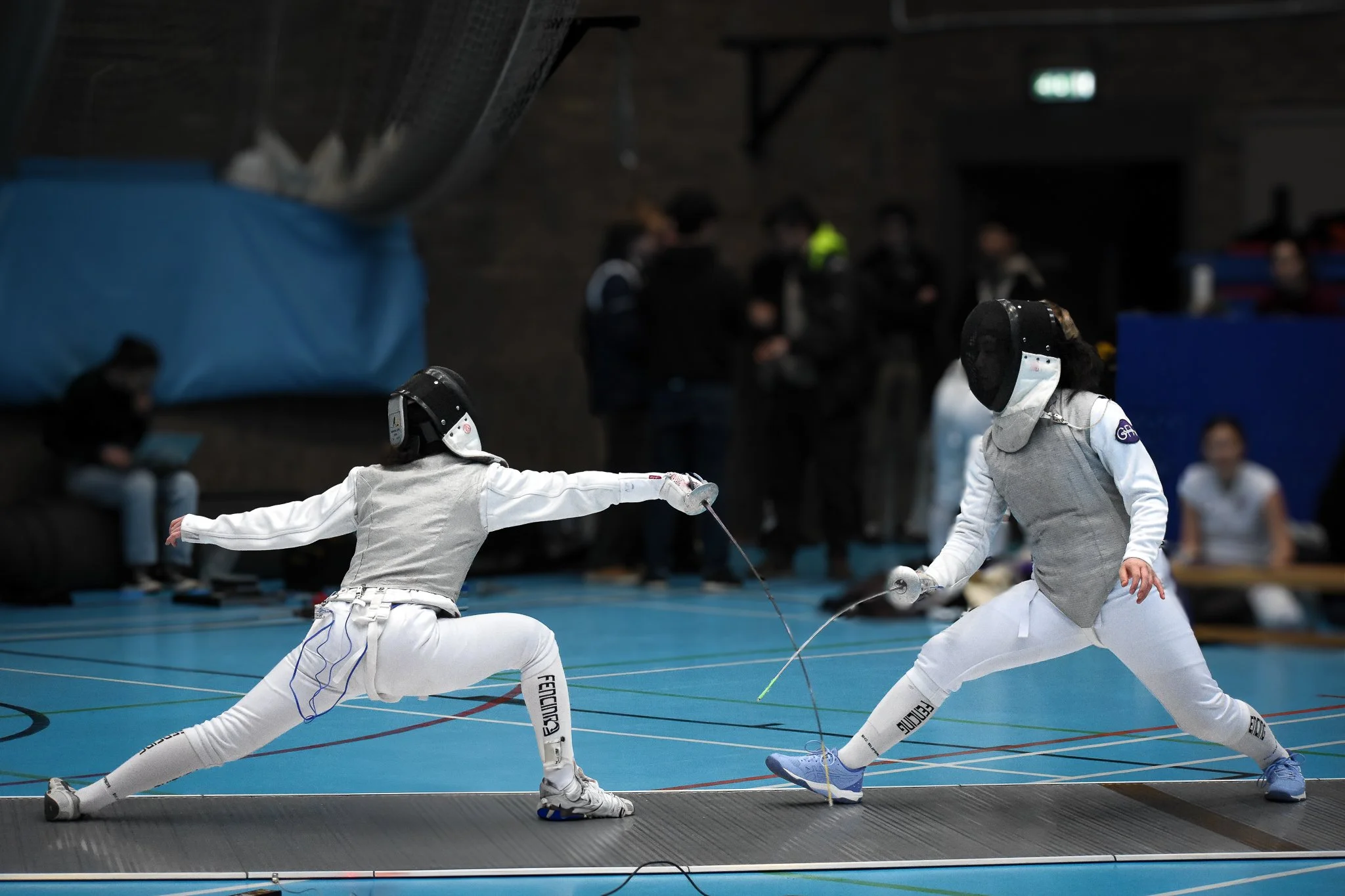 Two women in fencing gear engaging in a fencing match, lunging at each other with swords on an indoor gymnasium floor.