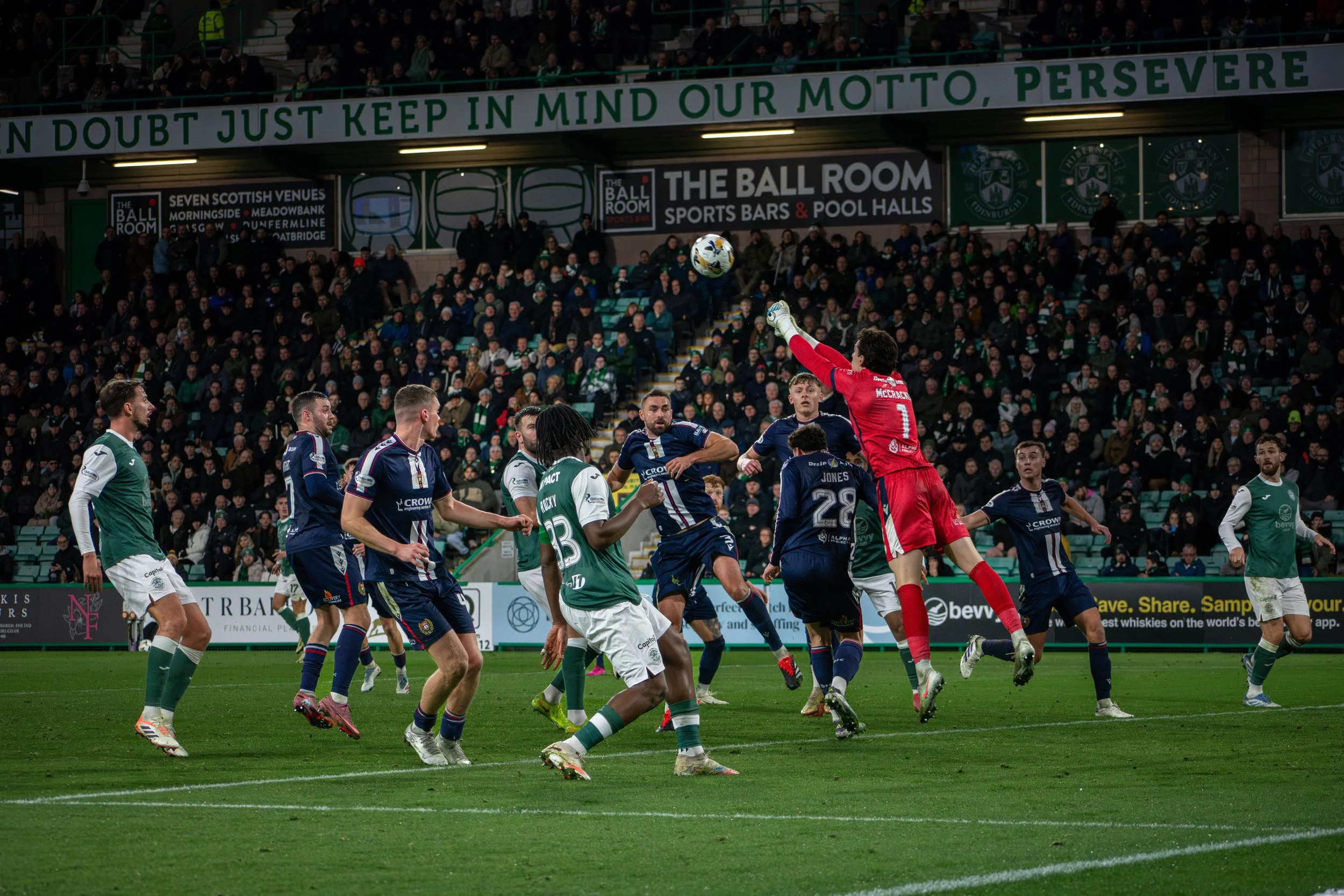 Soccer game with players jumping for a ball, goaltender in red uniform preparing to catch or block the ball, crowd in stadium stands, banners and advertisements in the background.