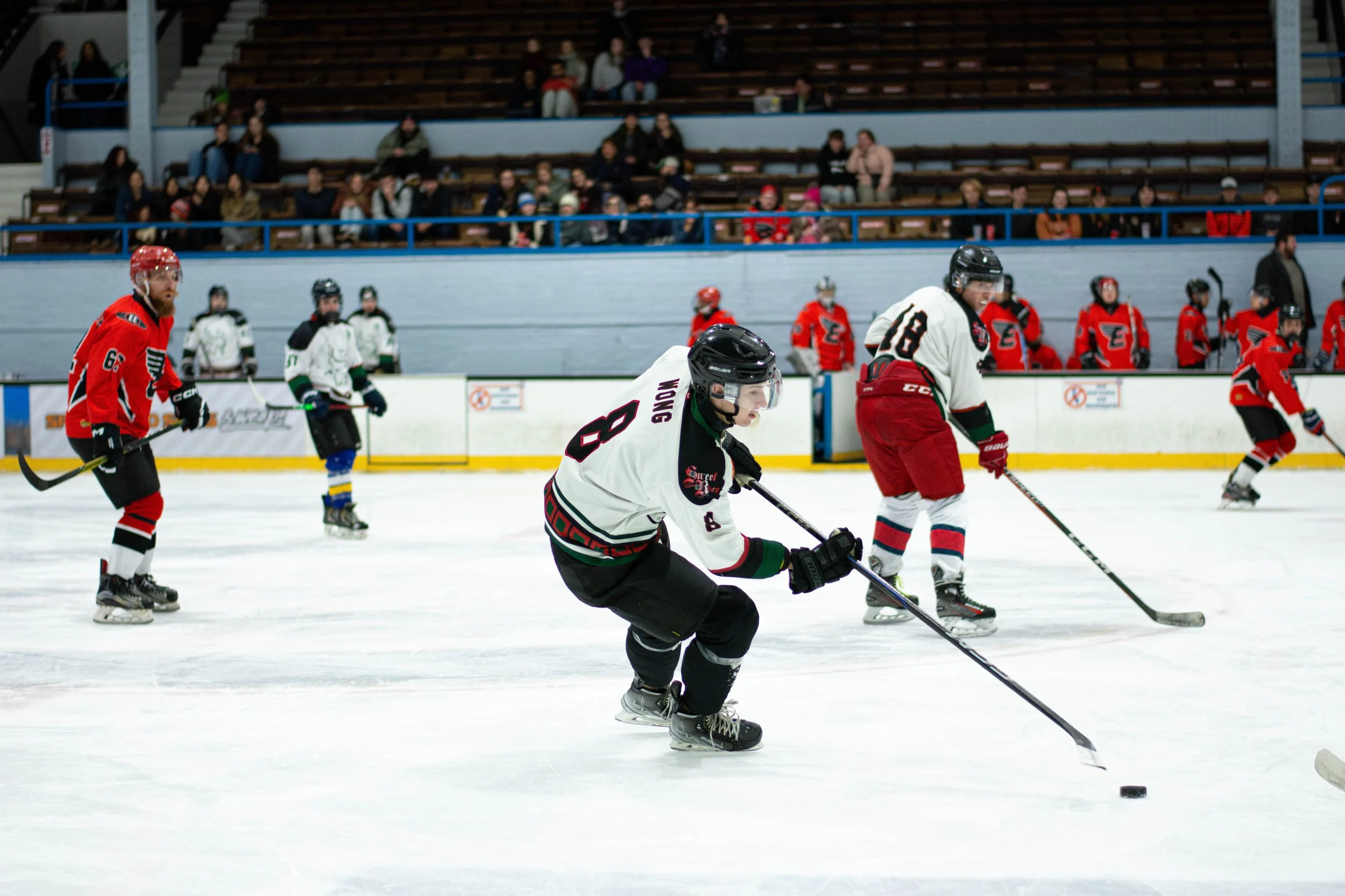 Hockey players in white and red jerseys on ice rink during game, with spectators watching from the stands.