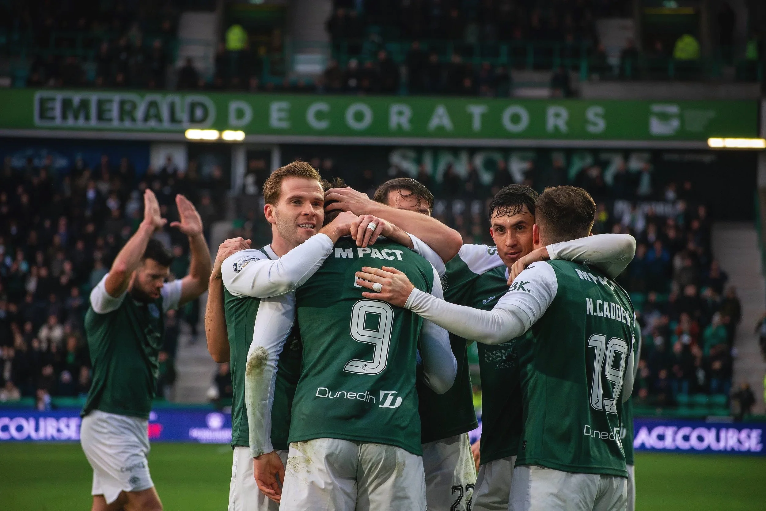 Players from Edinburgh's Hibernian FC celebrate together on the field, embracing each other in a huddle after a goal, with a crowd in the background.