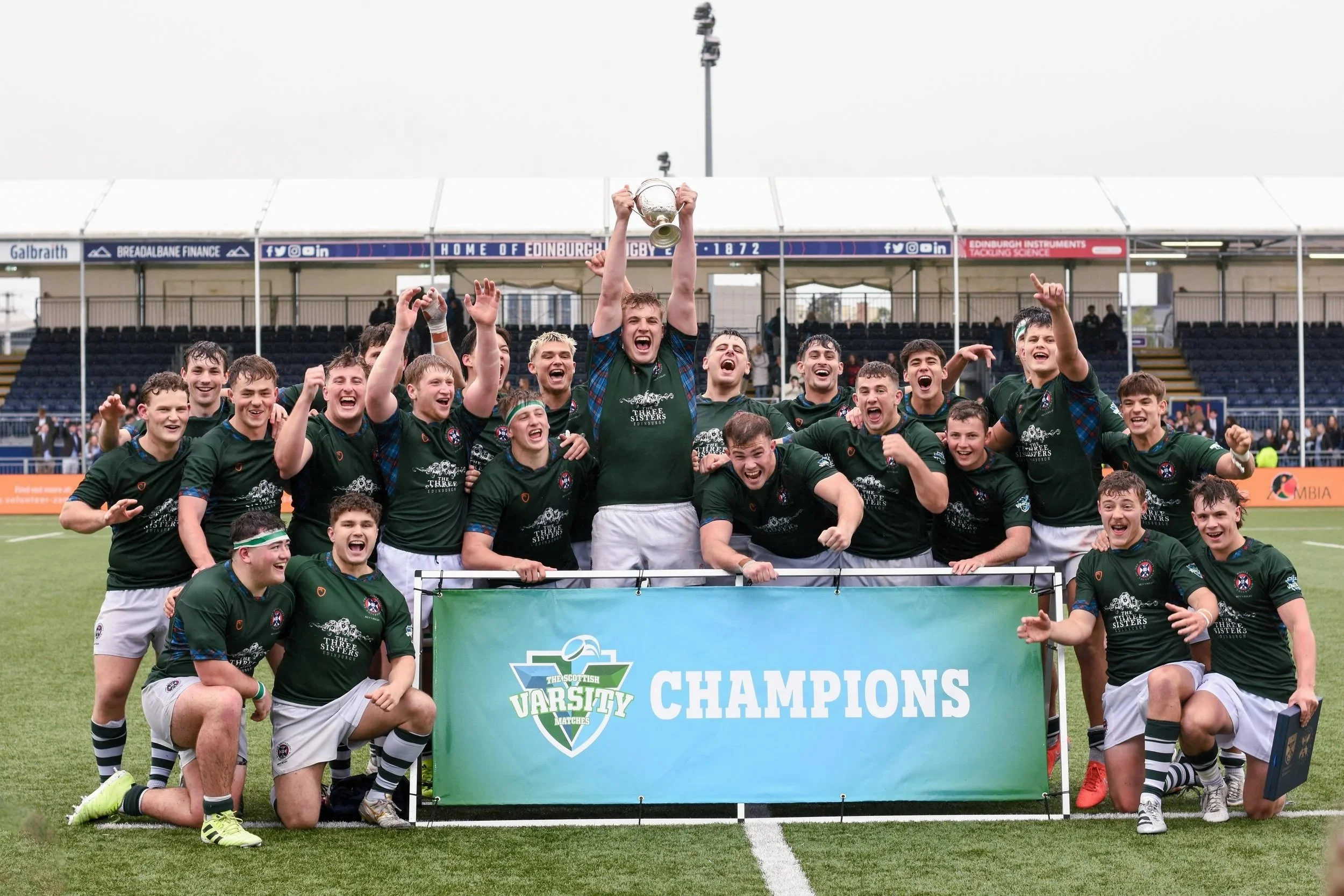 Edinburgh University's rugby team celebrating on the field after winning the 2025 varsity match as they hold a banner that reads 'CHAMPIONS' and the trophy, with players cheering and smiling.