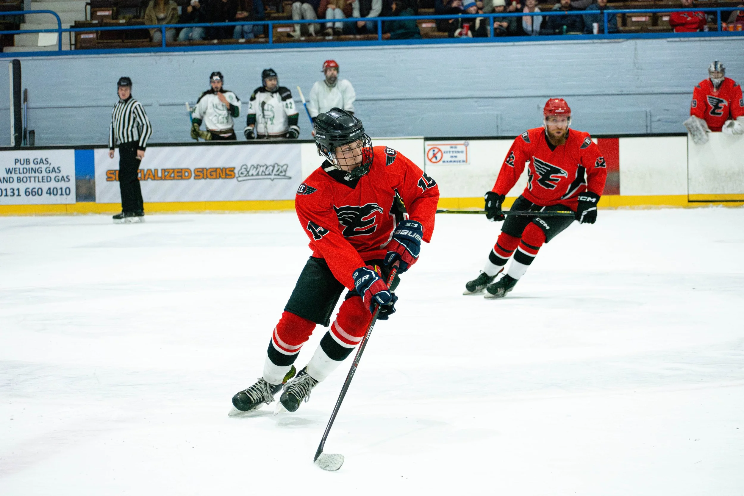 Ice hockey game with players in red uniforms, one controlling the puck, with spectators and other players in the background.