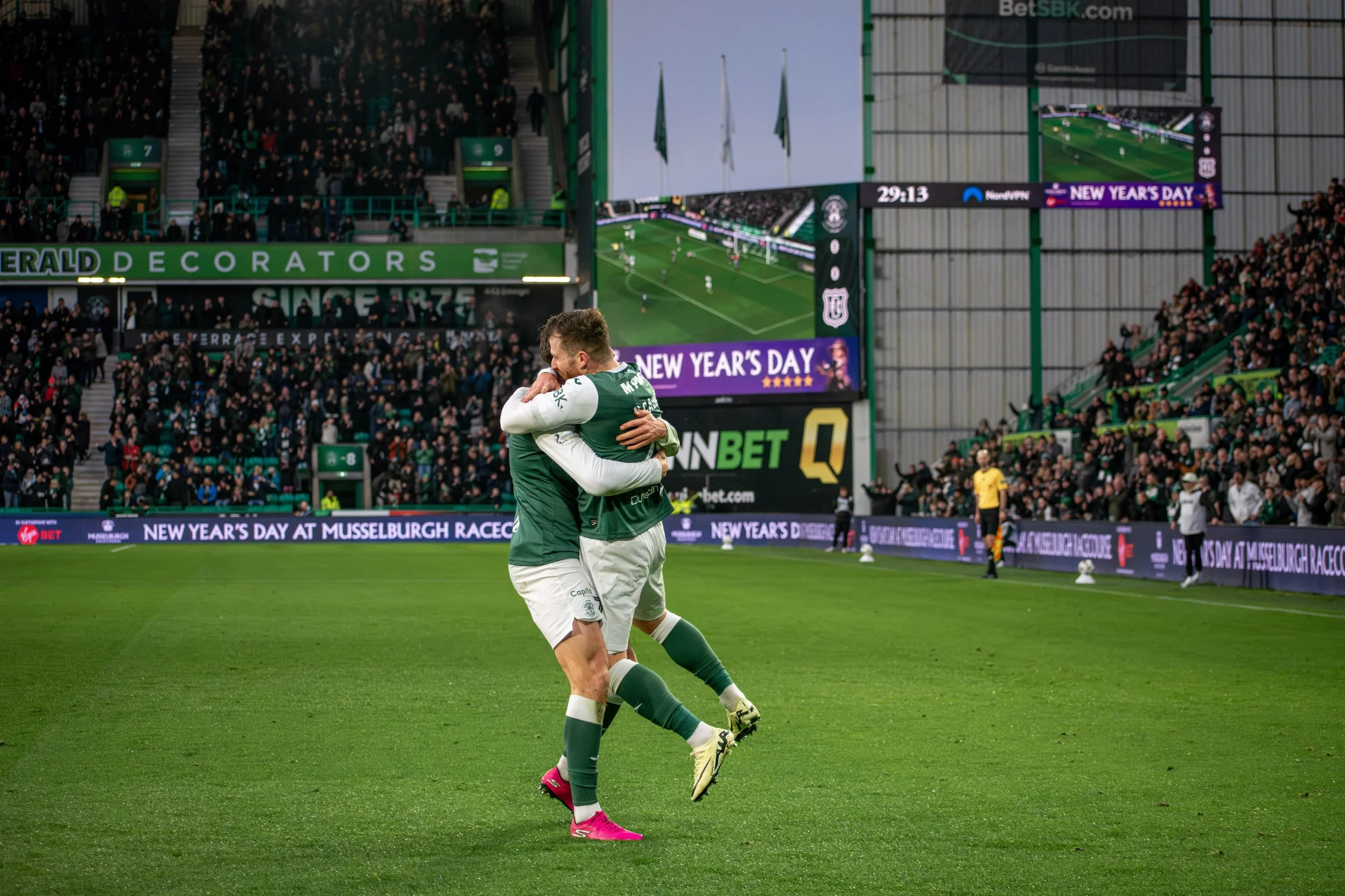 Two soccer players in green and white uniforms embrace and celebrate on the field, surrounded by a packed stadium with digital advertisements and screens, during a match at Musselburgh Racecourse.