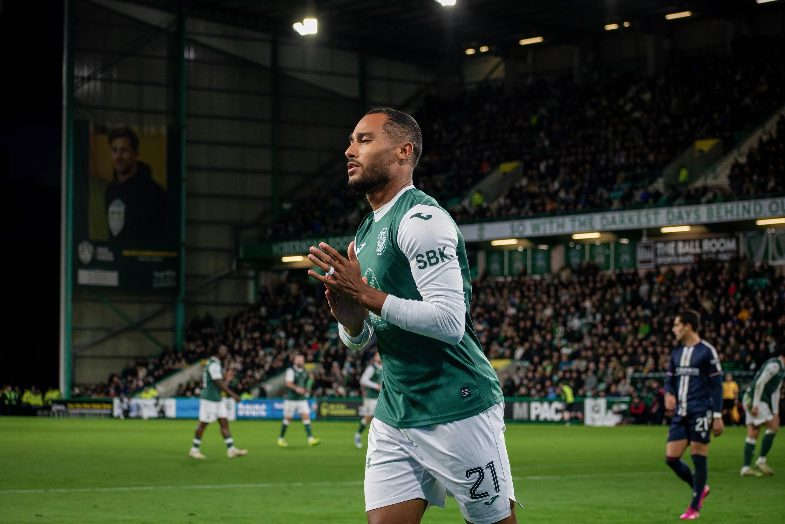 A soccer player in a green and white uniform with the number 21 is standing on the field with his hands clasped together, looking thoughtful. The stadium is filled with spectators. There are other players and a large banner in the background.