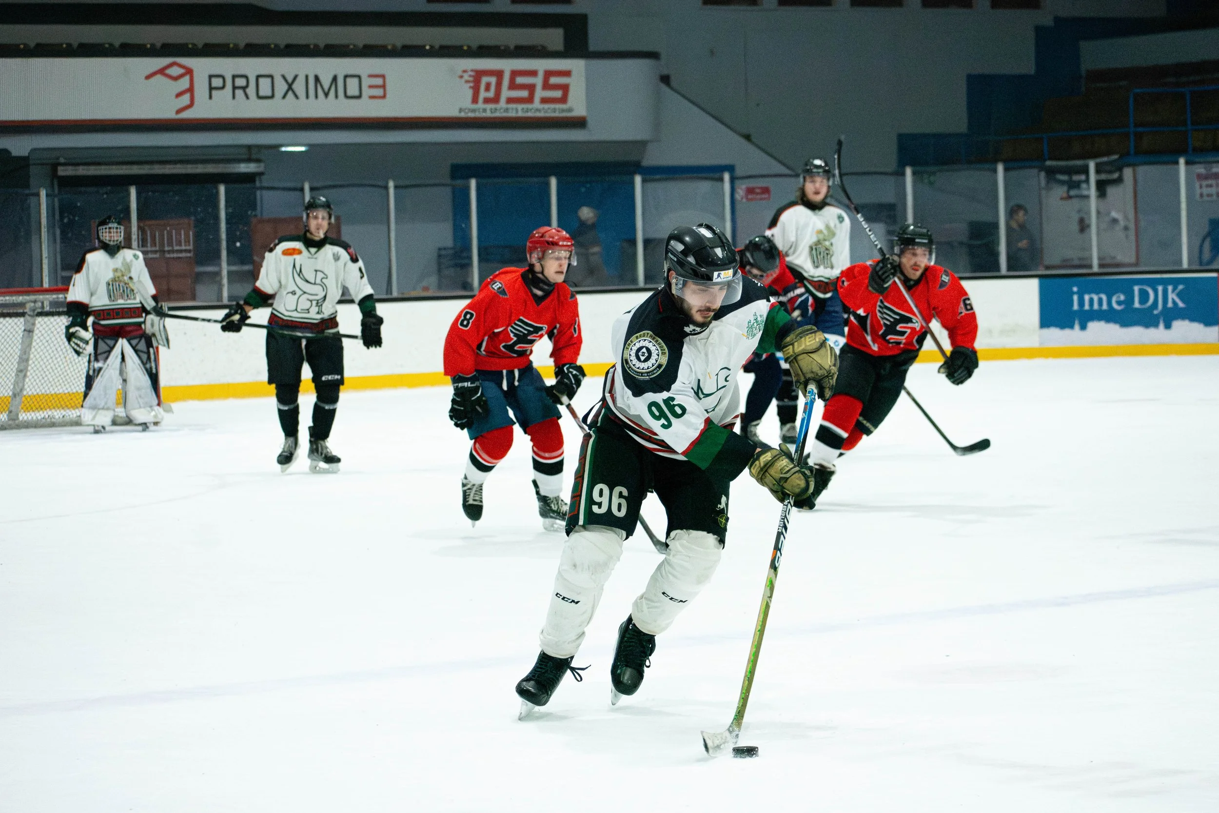 Hockey players on ice rink during game, including player in foreground wearing white jersey and black helmet, chasing puck, with other players in red and white jerseys in background.