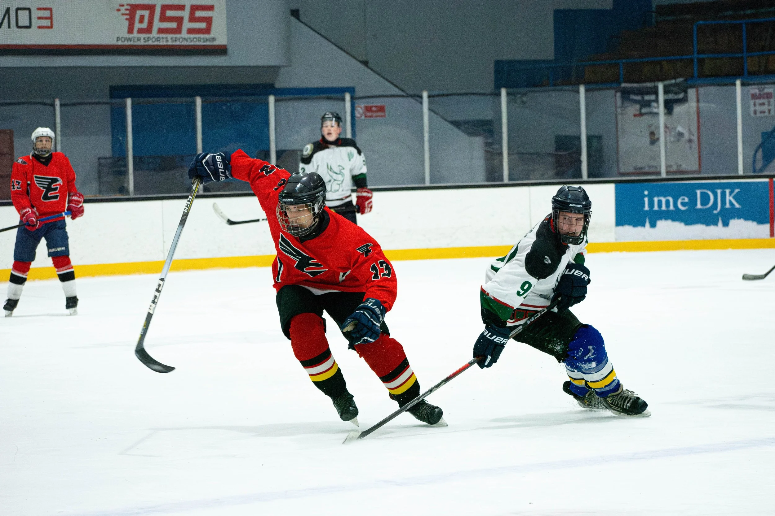 Hockey players in action on the ice rink during a game, with one player in a red jersey and another in a white jersey competing for the puck.