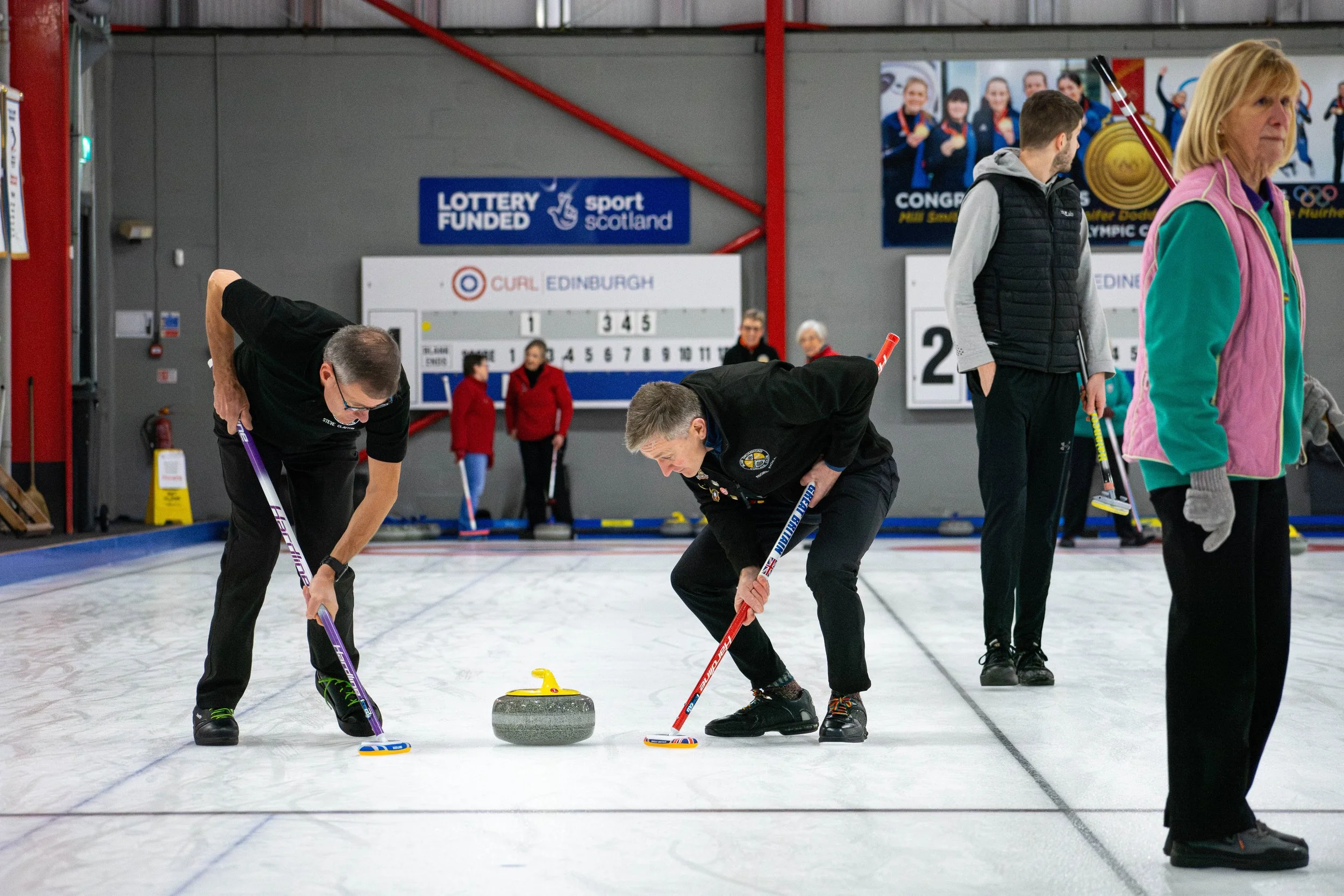 Two men are curling on an indoor ice rink, preparing to release a curling stone while other players and officials observe in the background.