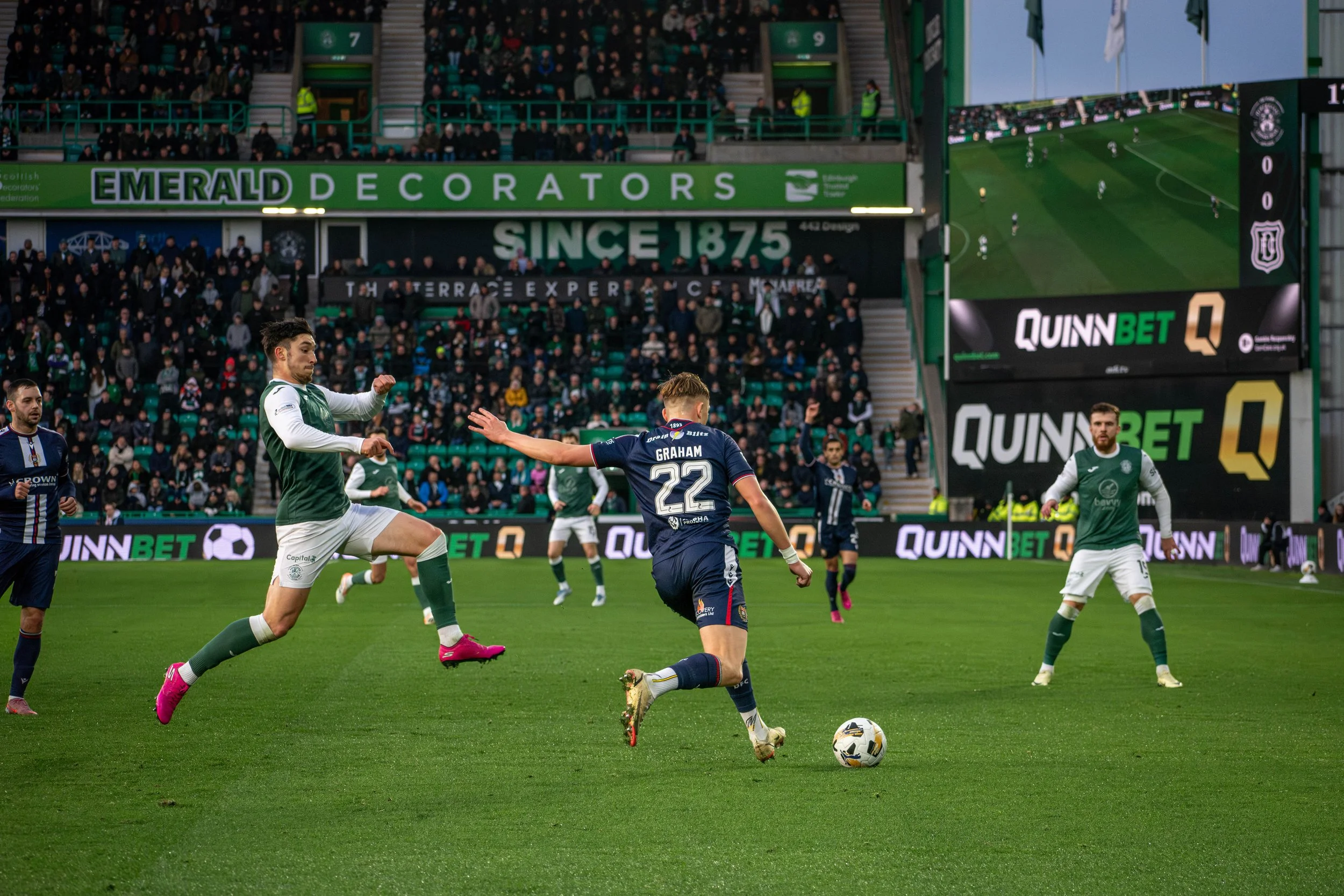 Soccer players on the field during a match. One player in a navy uniform is kicking the ball, while another in a green and white kit is jumping to block. The crowd is seated in the stands in the background.