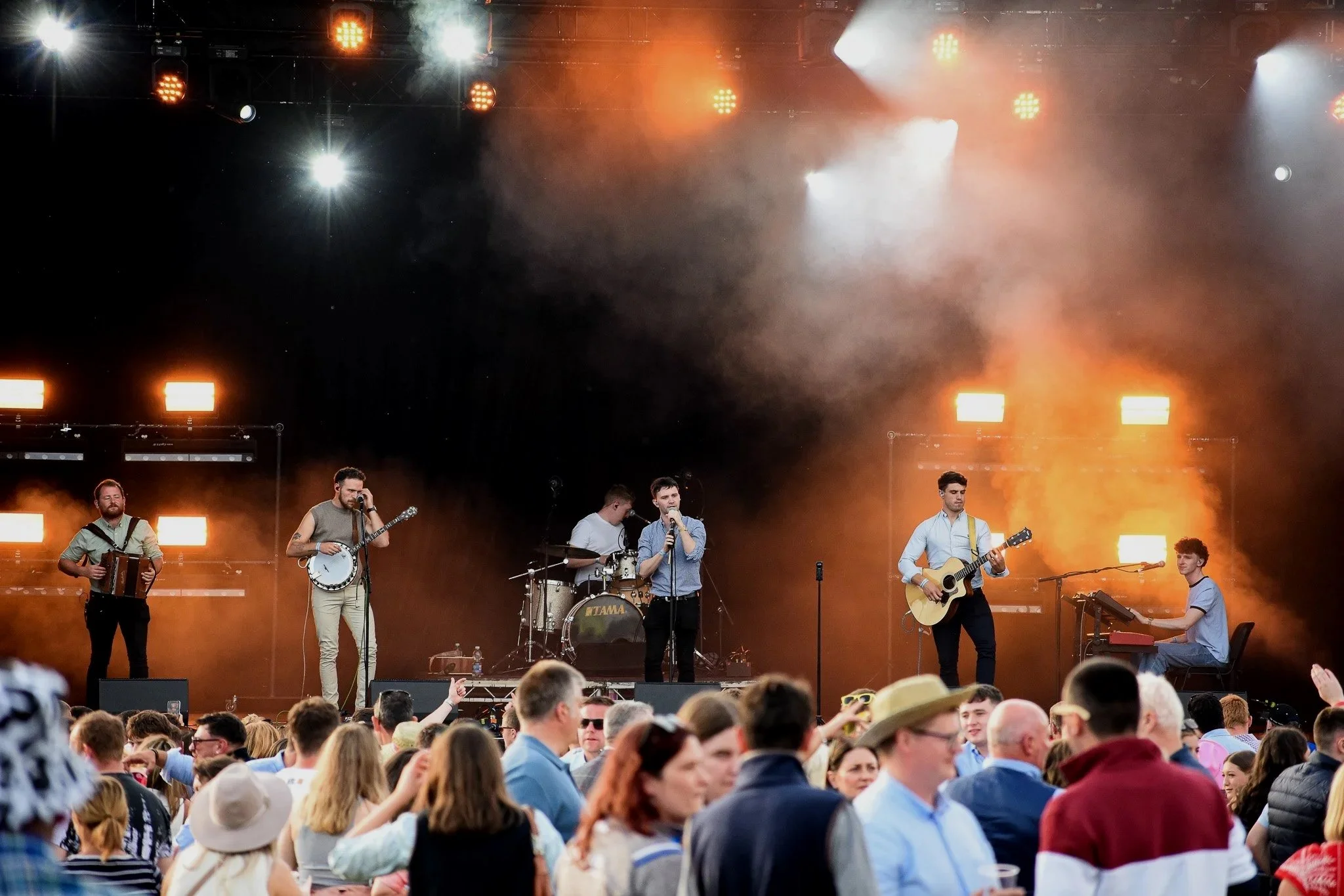 A band performs on stage during a concert with orange and white lighting and a large crowd watching in front.