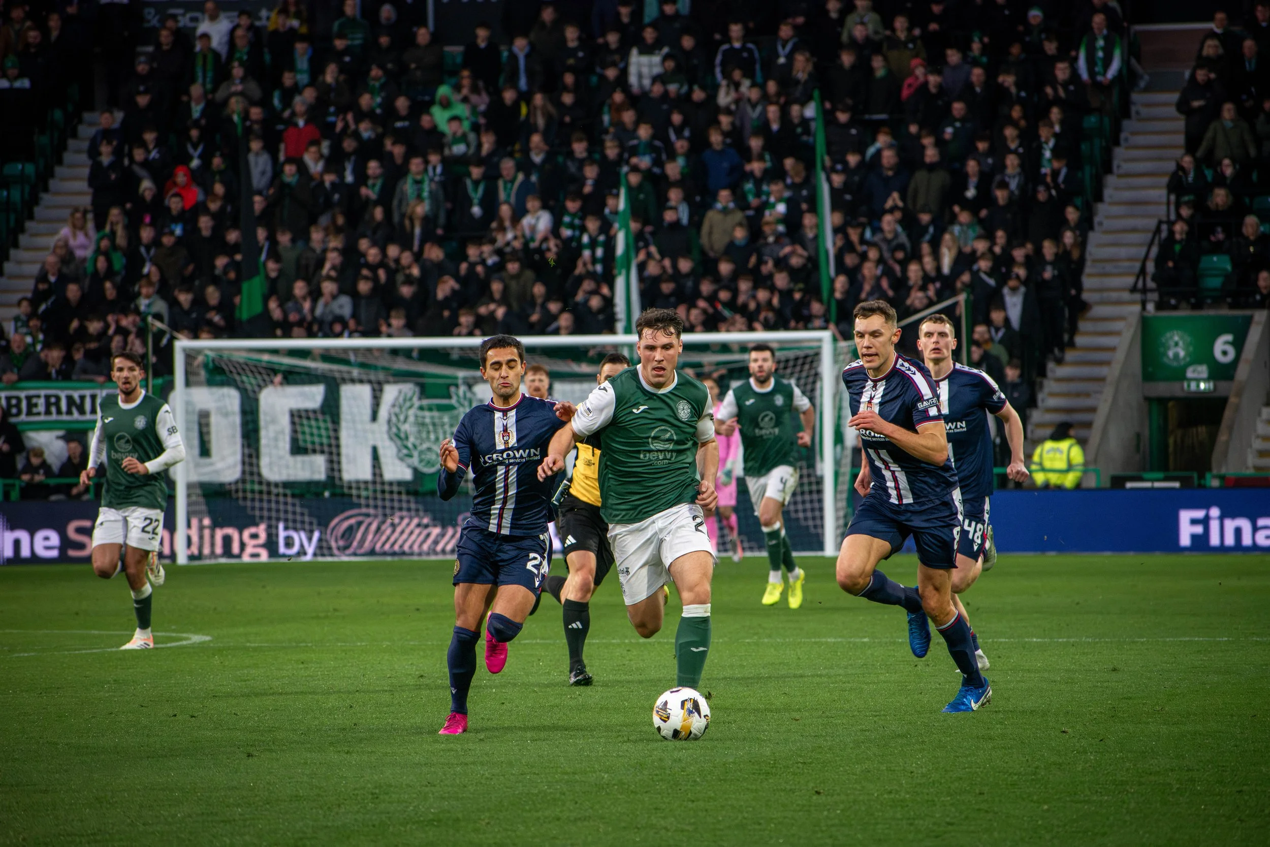 Soccer players in action during a match, with a green team player in the center kicking the ball, near the goal, surrounded by players in dark blue jerseys and a large crowd in the stands.