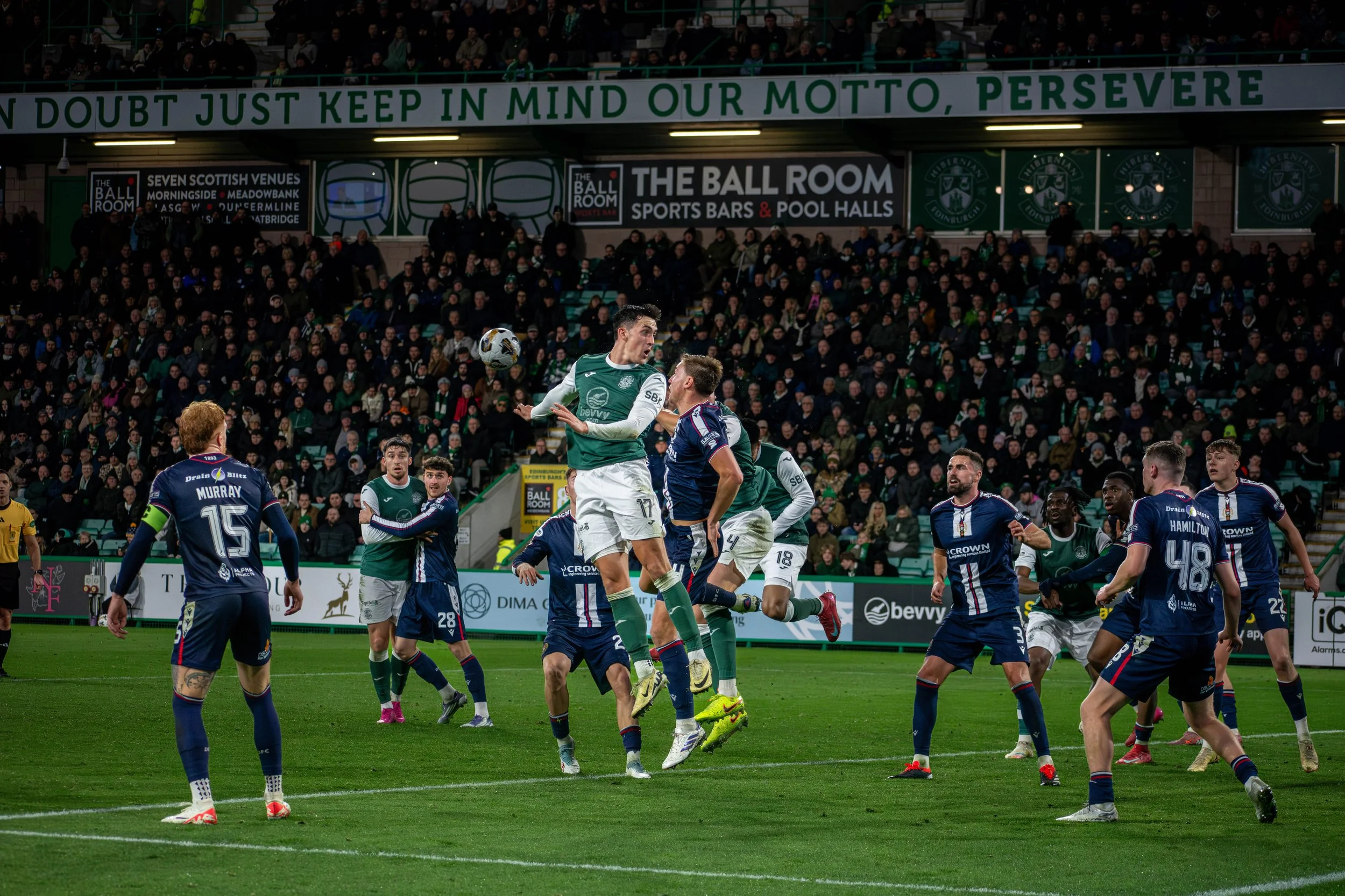 Soccer match with players jumping for the ball, some players wearing green and white uniforms and others in navy blue and white kits, on a green field with a crowd in the stands.