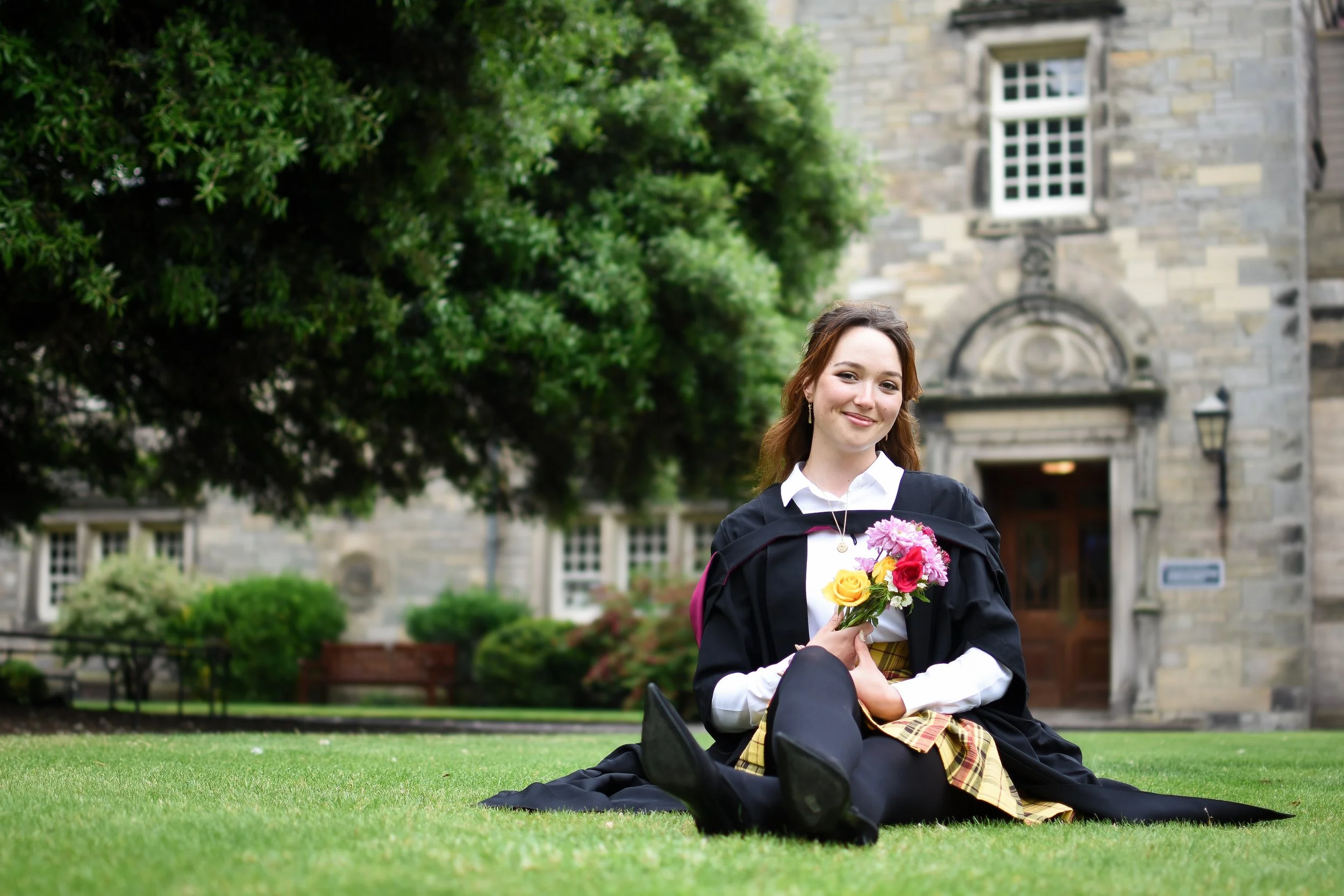 A young woman in graduation attire sitting on grass lawns outside a historic stone building, holding a bouquet of pink, yellow, and red flowers, smiling at the camera.