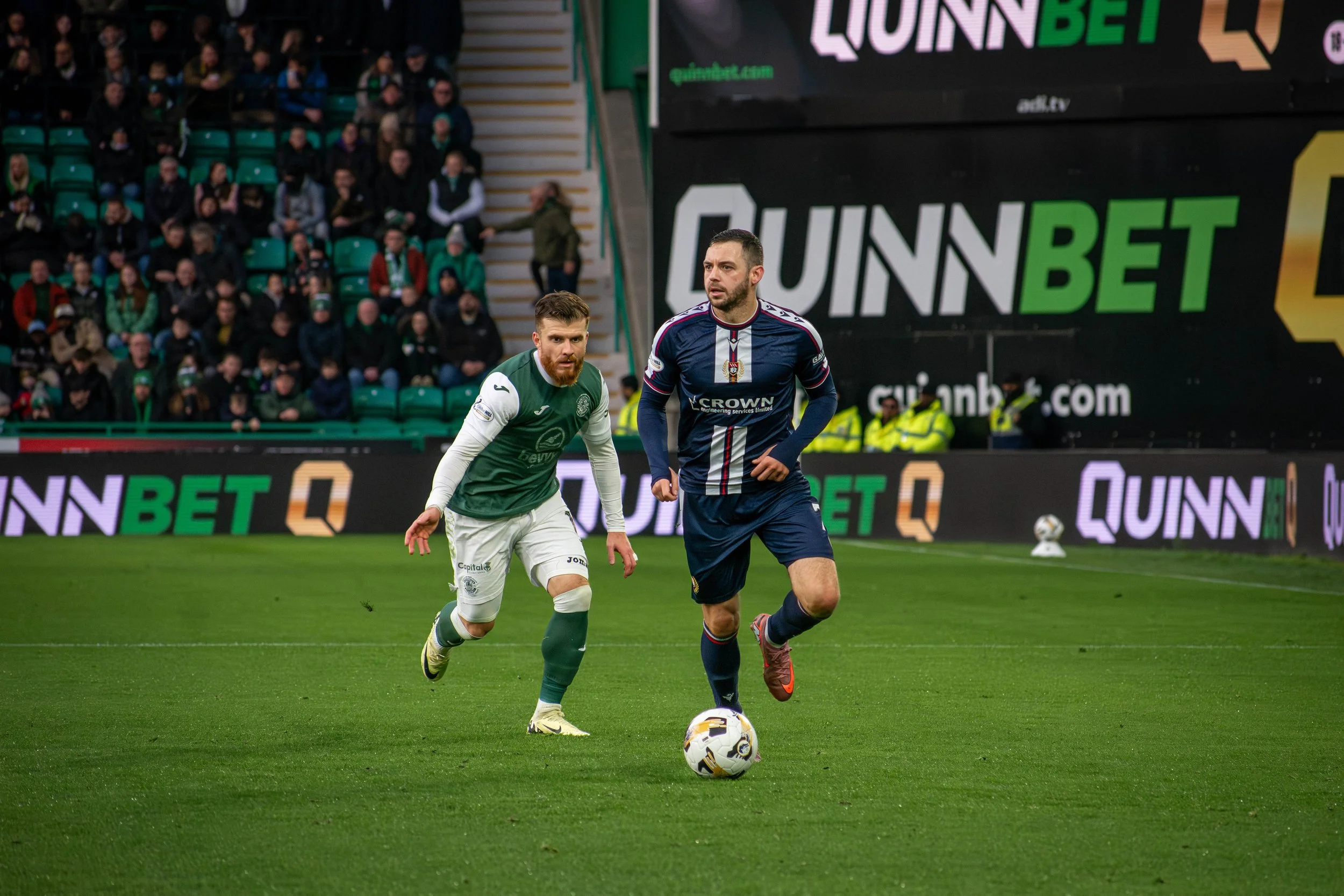 Two soccer players on a field during a match, one in a green and white uniform, the other in a blue and red uniform, with a crowd in the stands and advertising boards in the background.