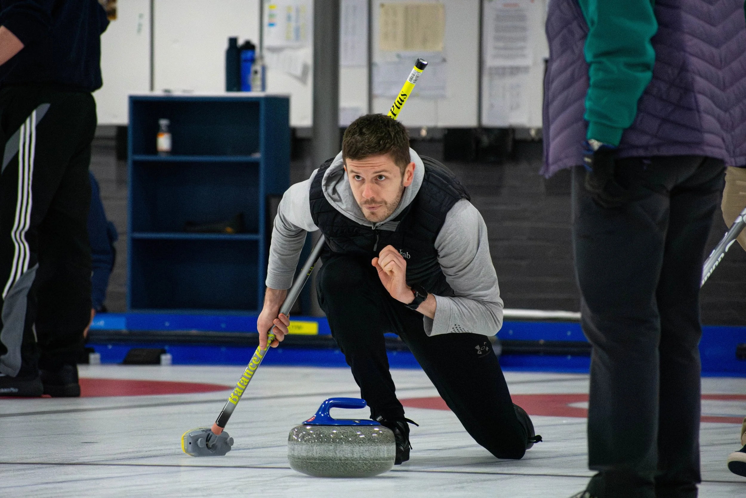 A man participates in a curling match, crouched on one knee on the ice, holding a broom, and preparing to release a curling stone.
