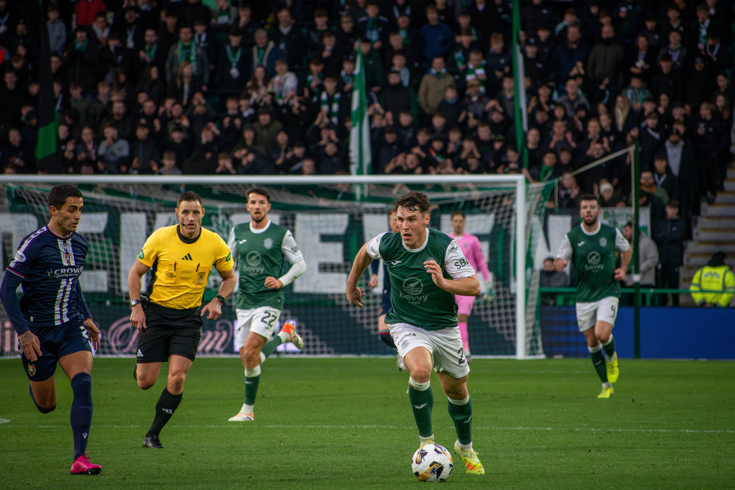 A soccer match in progress with players running on the field. One player in a green jersey is controlling the ball, while other players and the referee are nearby. The audience is visible in the background.