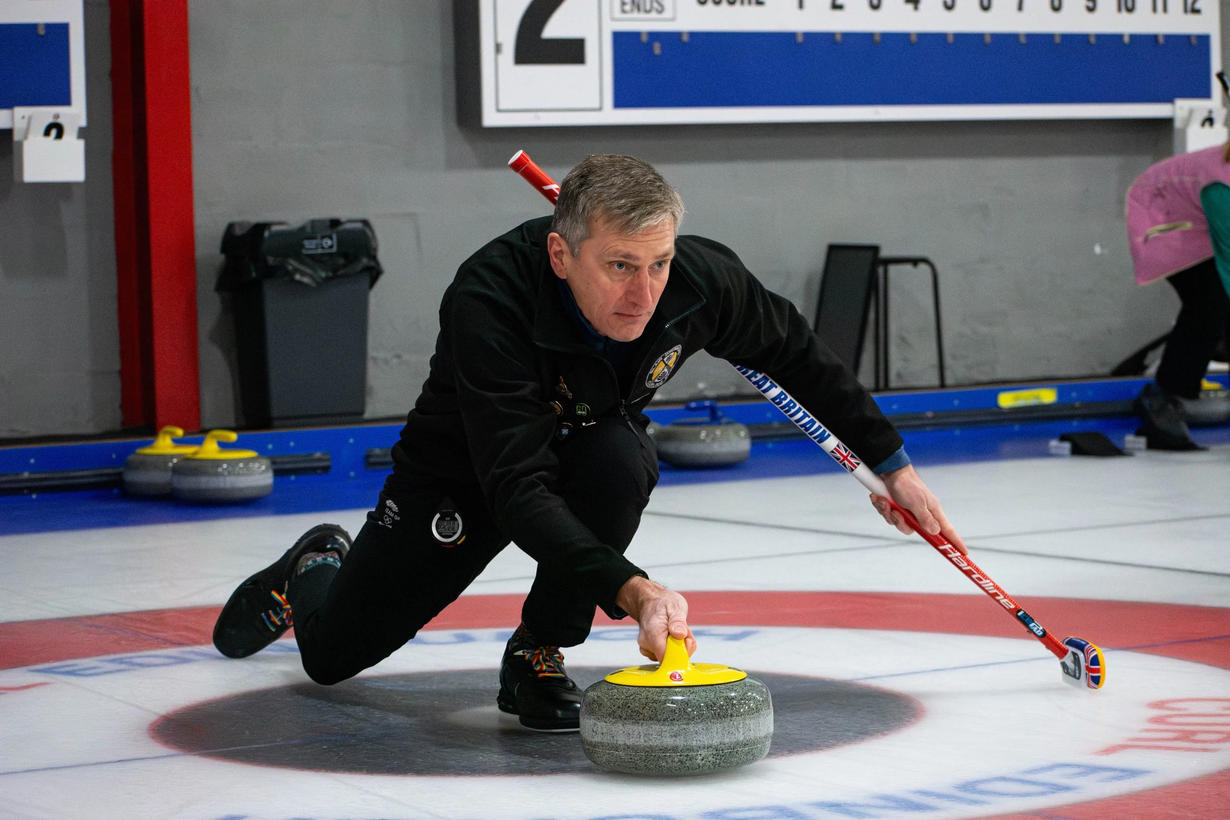 A male curling player in black attire is delivering a curling stone on an indoor ice rink, with other players and curling equipment visible in the background.