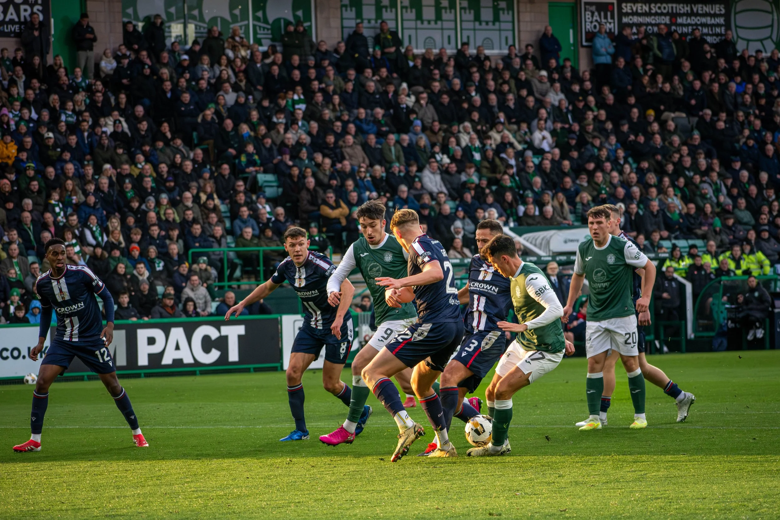 Soccer players on a field competing for the ball during a match, with a large crowd in the stands.