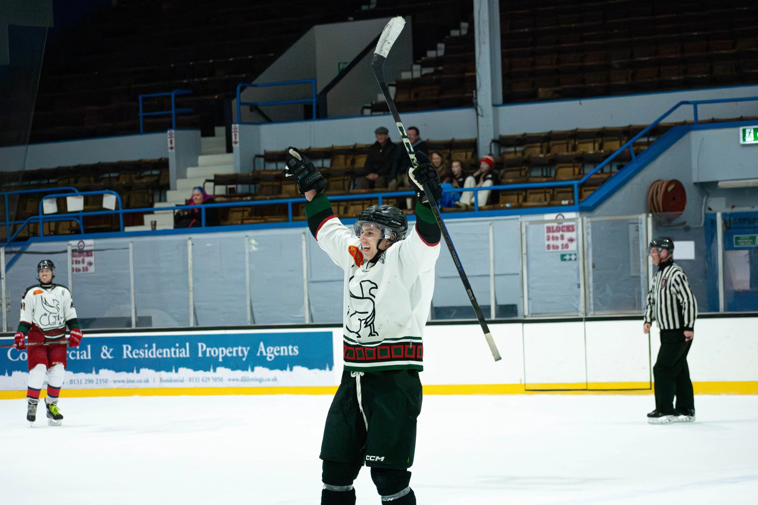 Ice hockey player celebrating on the ice rink with raised arms, holding a hockey stick, wearing a white jersey with black and red accents, black helmet, and black gloves; other players and a referee are visible in the background, along with empty sta