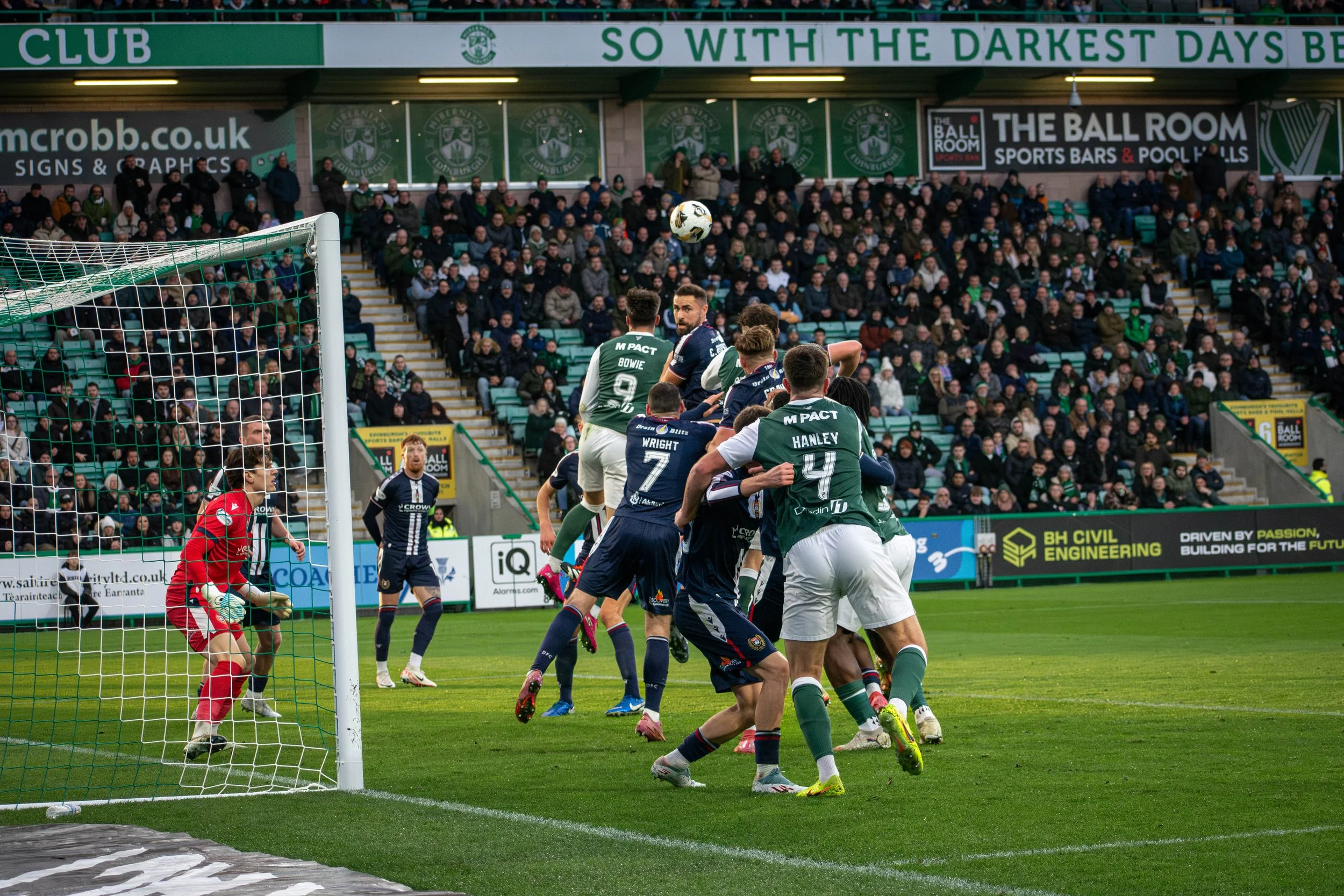 Soccer players from two teams leap and jump in front of the goal during a match, with spectators watching from the stands. The ball is mid-air near the players, and the goalkeeper is prepared on the left.