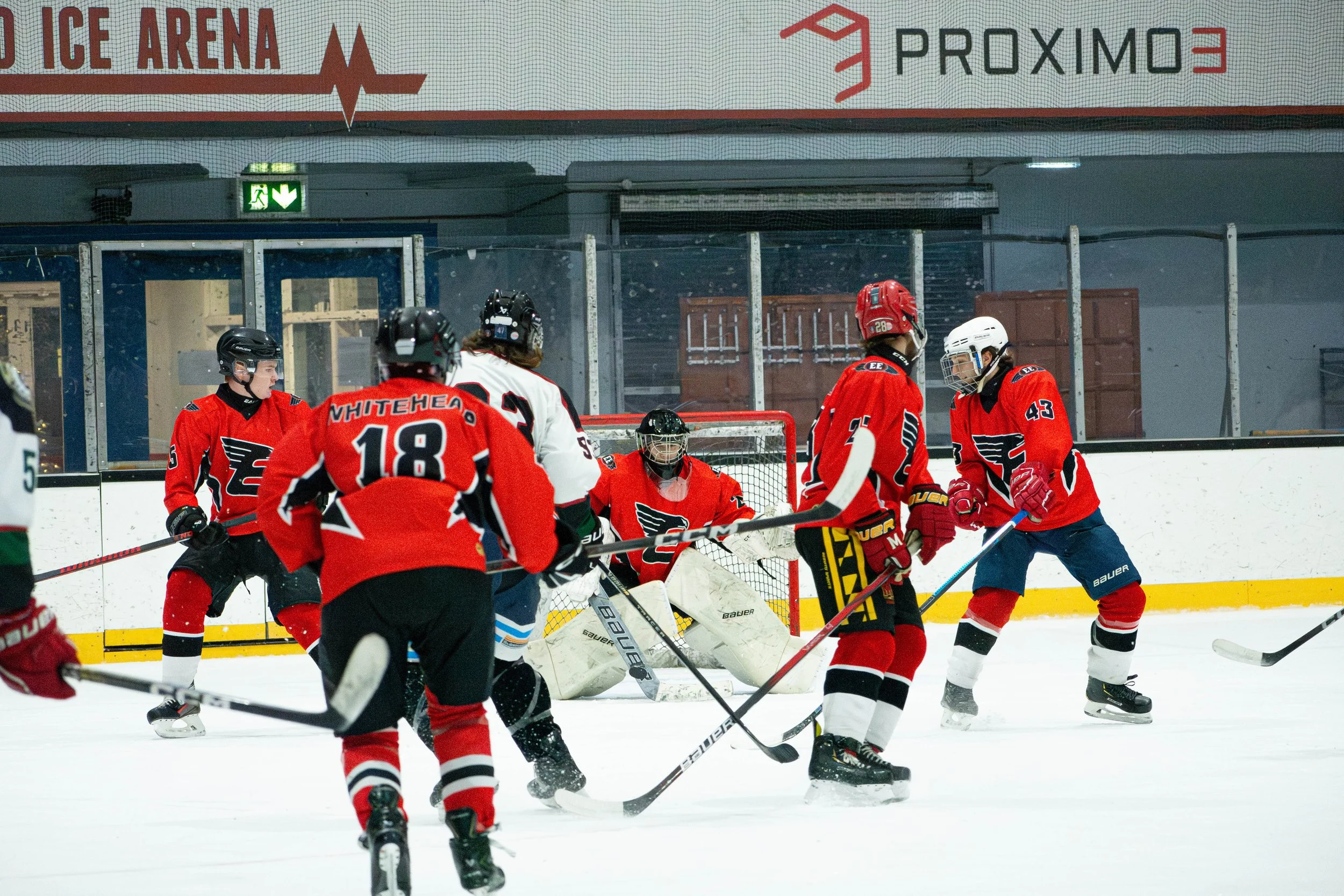 Hockey players on ice rink in a game, with some players skating and one goalie in front of the net, all wearing red jerseys with black and white accents.