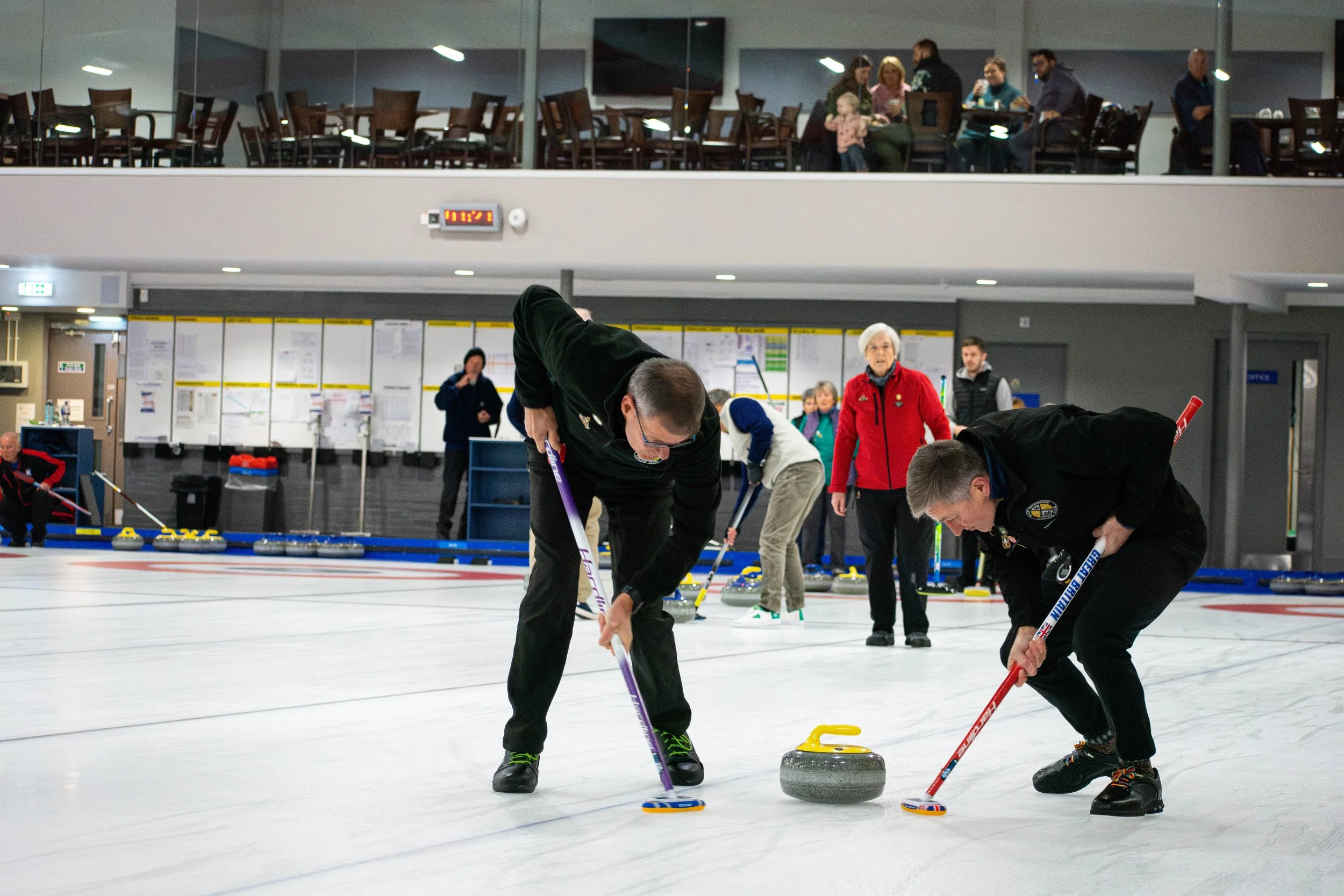 Two men playing curling on an indoor ice rink, with others and spectators in the background.