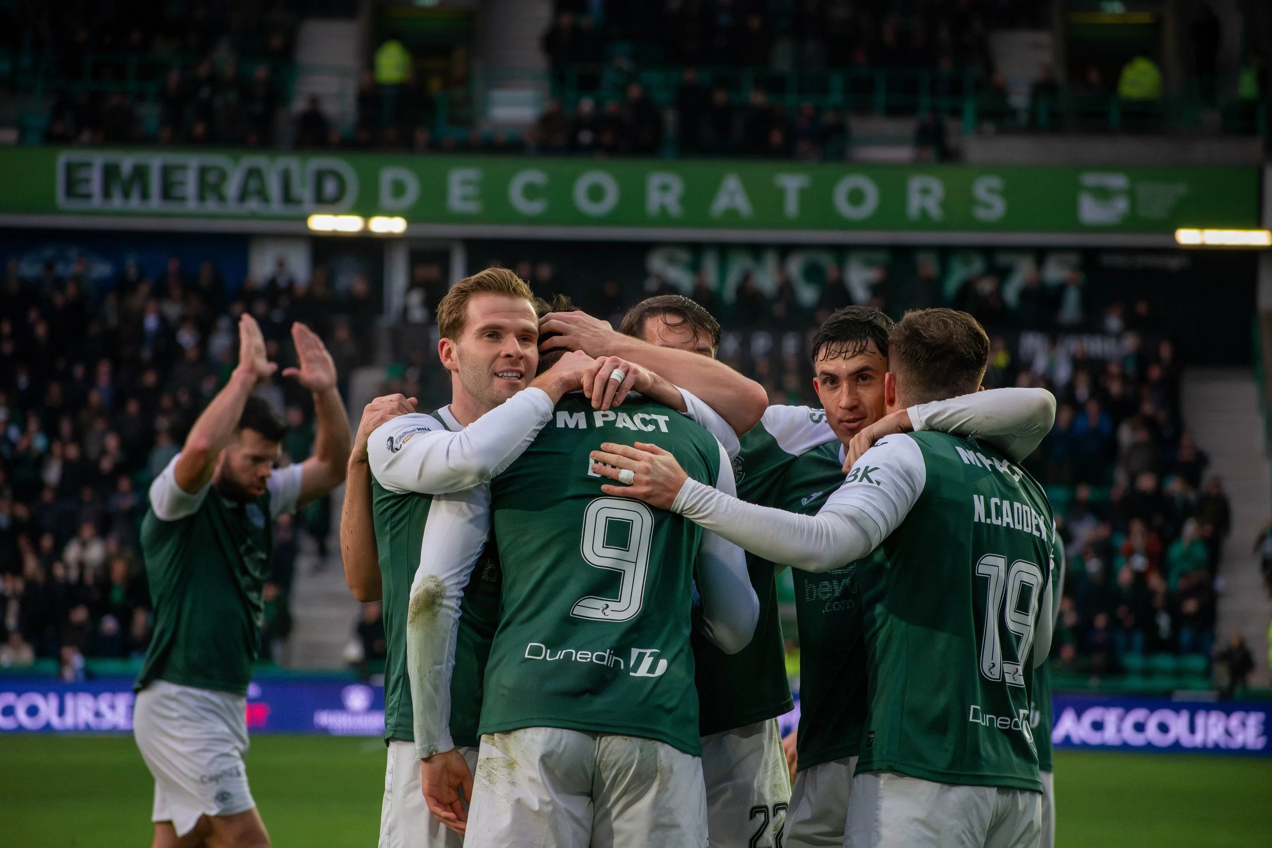 Soccer players in green jerseys celebrating on the field with one player being lifted in a group hug, and some fans in the background.