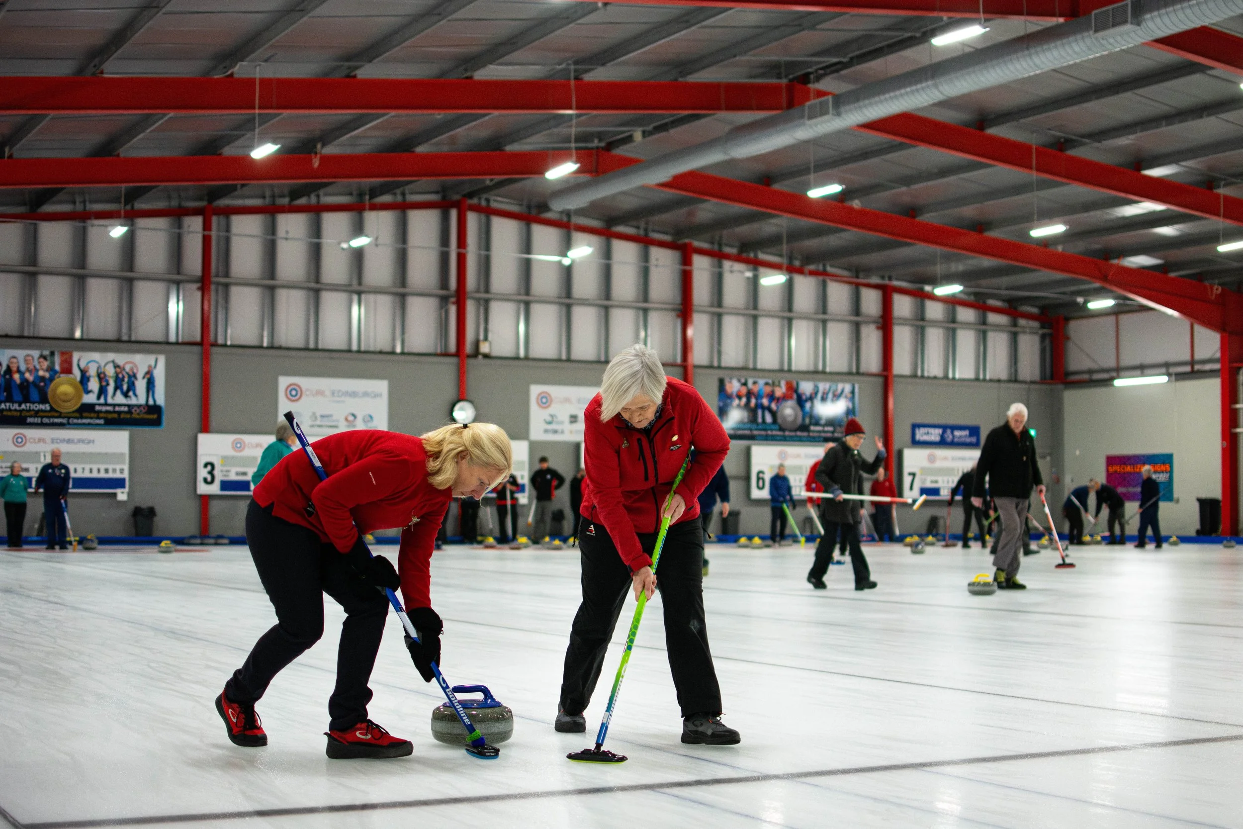 People participating in an indoor curling game, some sweeping the ice near the curling stones, with other players in the background on the ice rink.