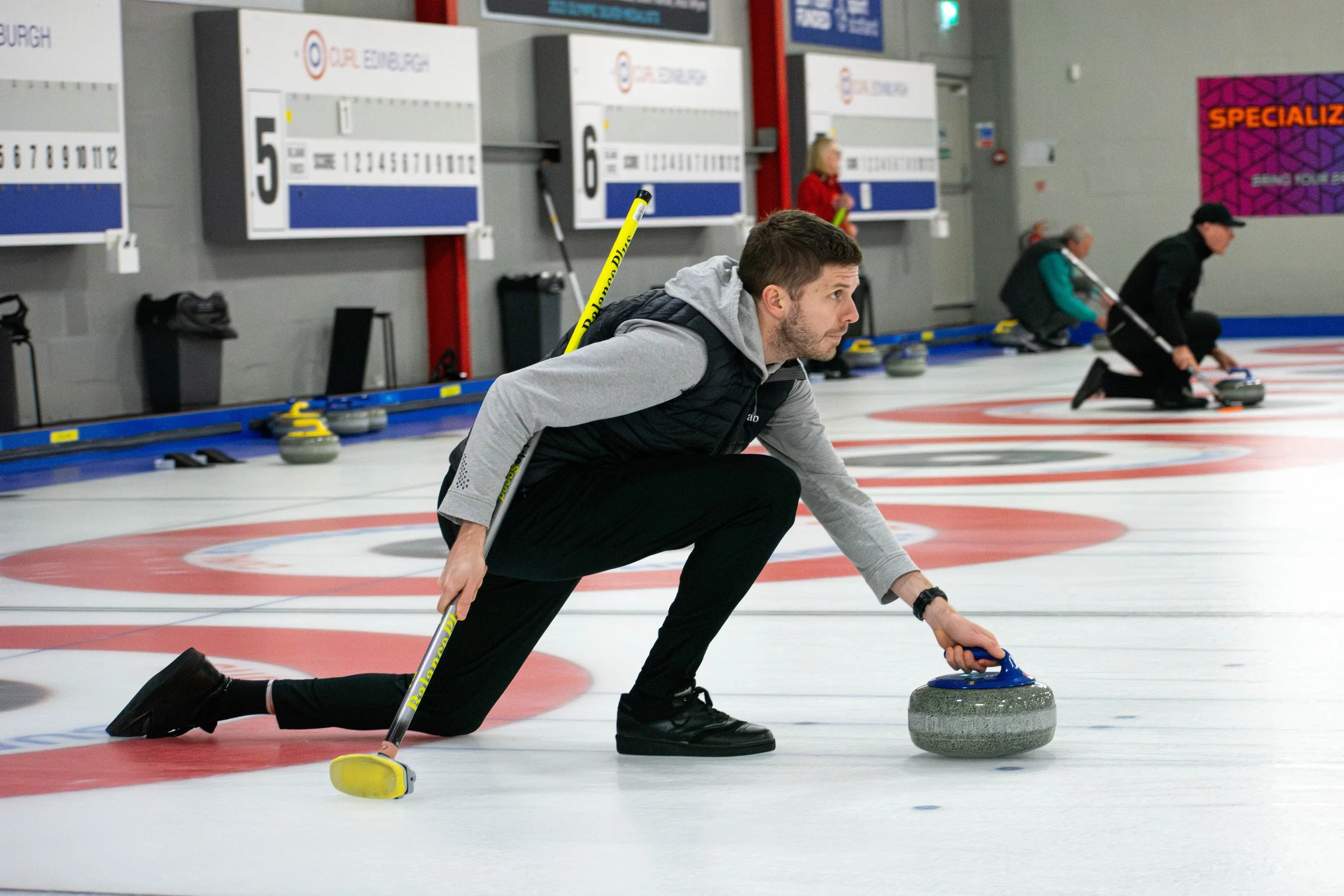 A man playing curling on an indoor ice rink, dressed in a gray and black jacket and black pants, is holding a curling stone in one hand and a broom in the other, preparing to slide the stone on the ice while kneeling on one knee.