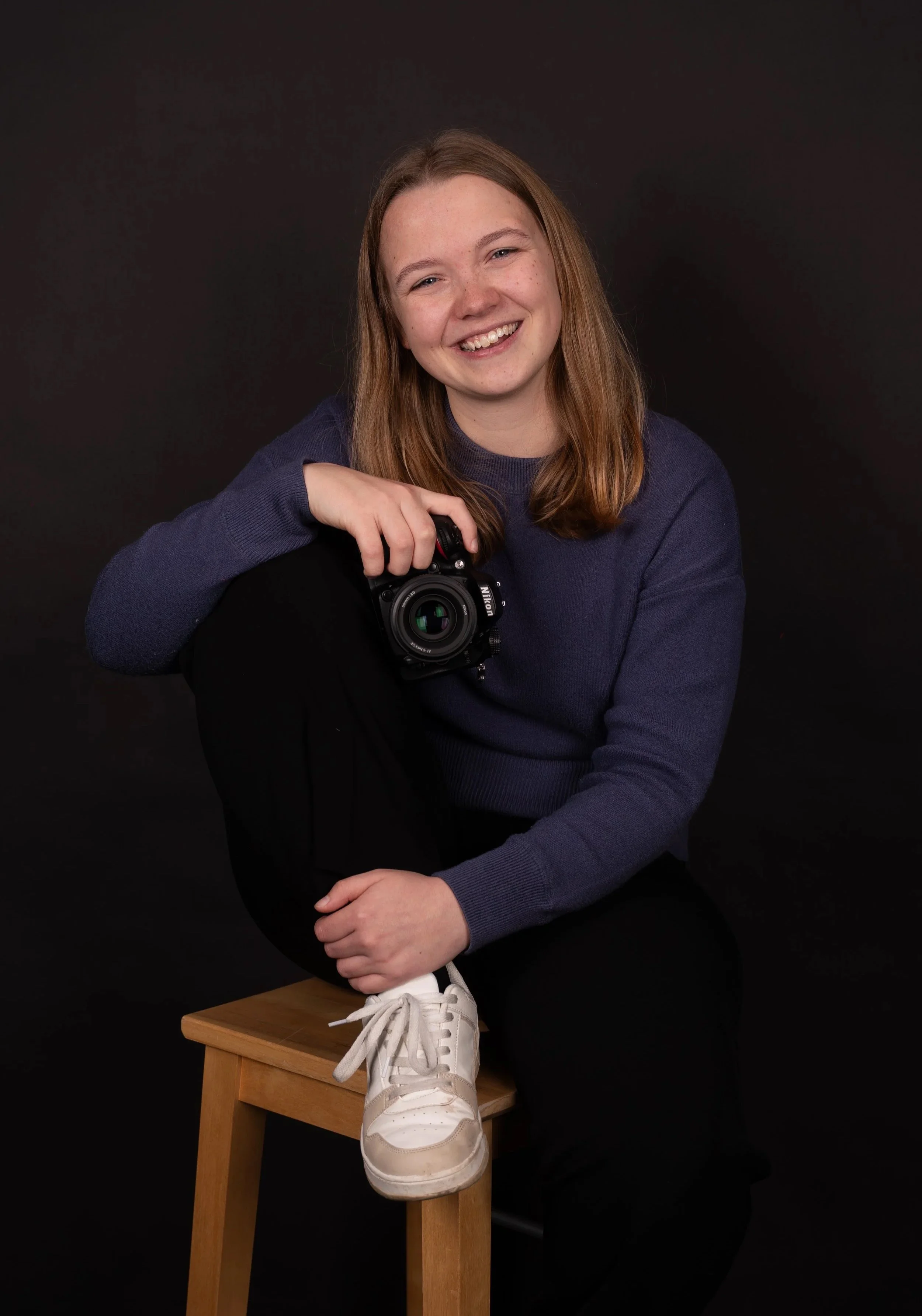 Young woman with blue hair, wearing a blue sweater, sitting on a wooden stool, holding a camera, and smiling against a dark background.