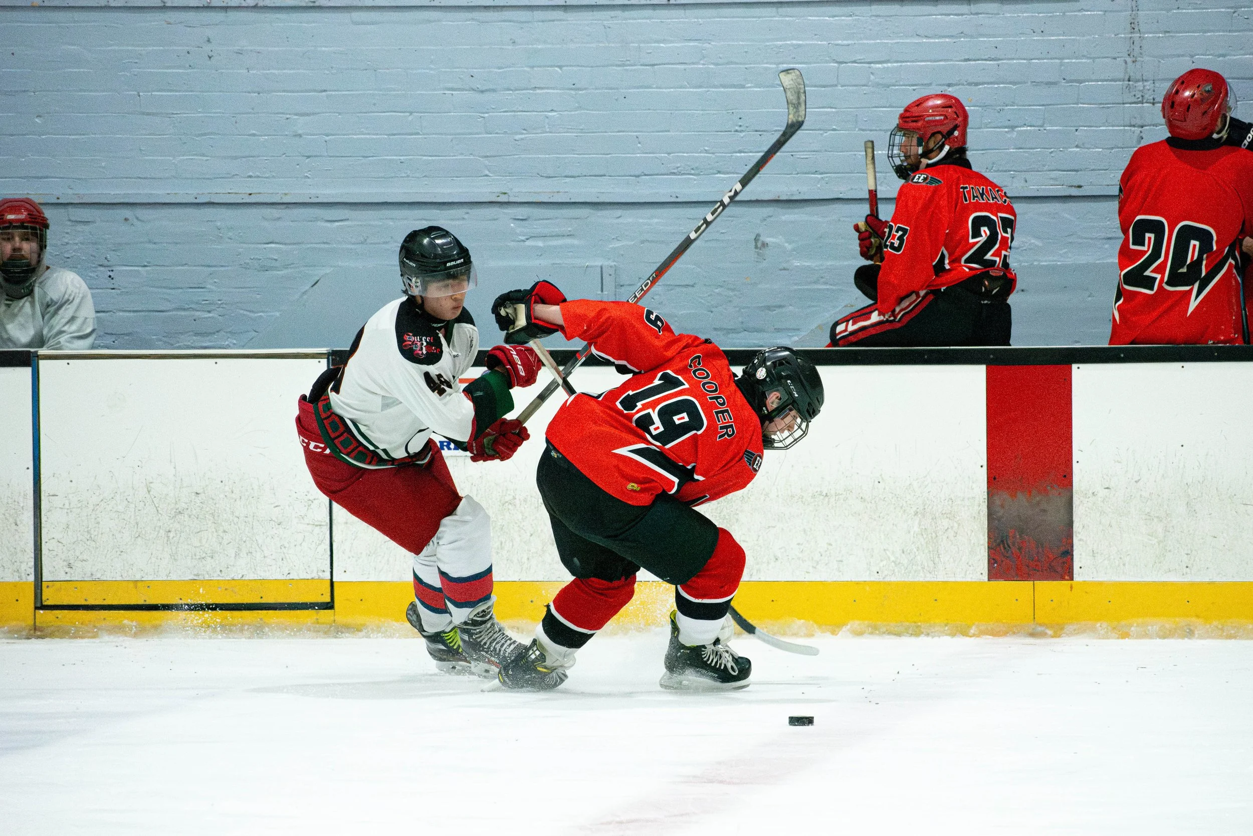 Two young hockey players, one in a white jersey and the other in a red jersey, compete for control of a puck on the ice rink. The player in red is leaning forward while the player in white reaches for the puck with their stick. In the background, thr