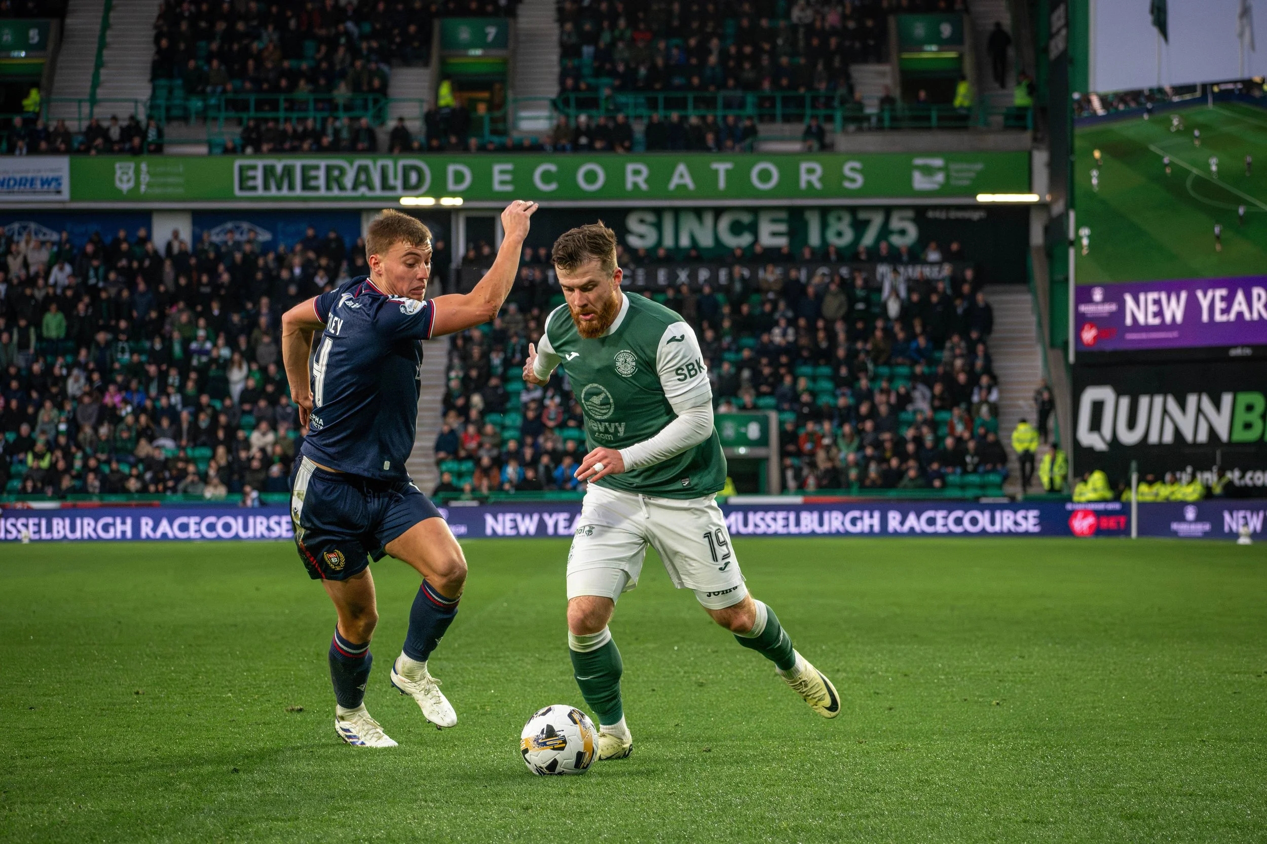 Two soccer players competing for the ball on a field at a stadium during a match, with a crowd in the background.