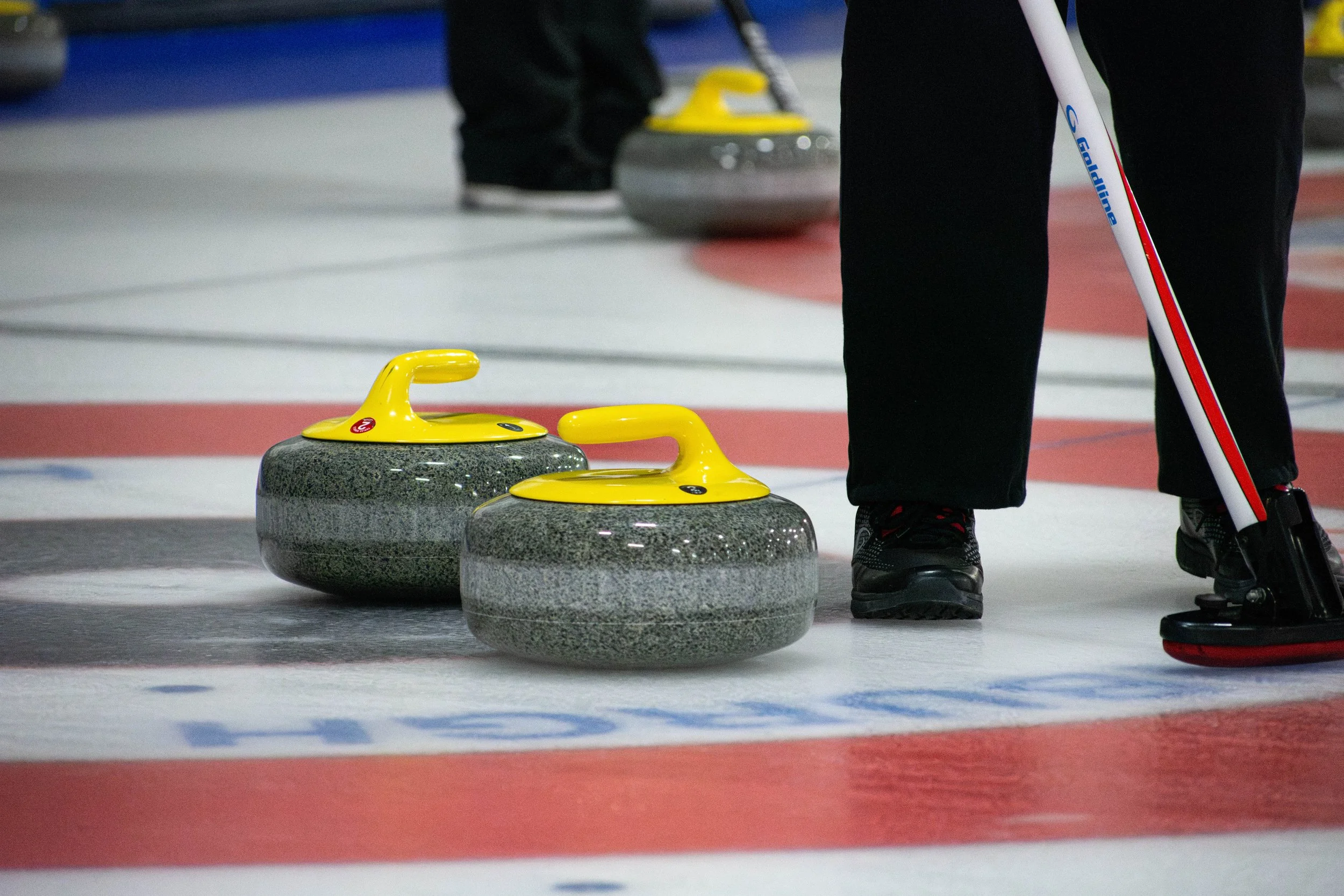 Two curling stones with yellow handles on an ice rink, with a person holding a curling broom nearby.
