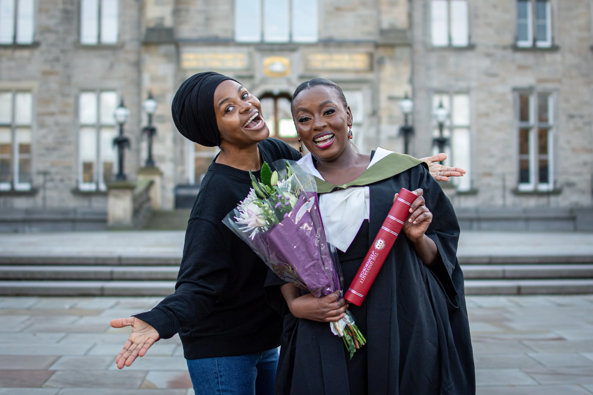 Two women celebrating graduation outside a university building, one holding a bouquet of flowers and the other holding a diploma