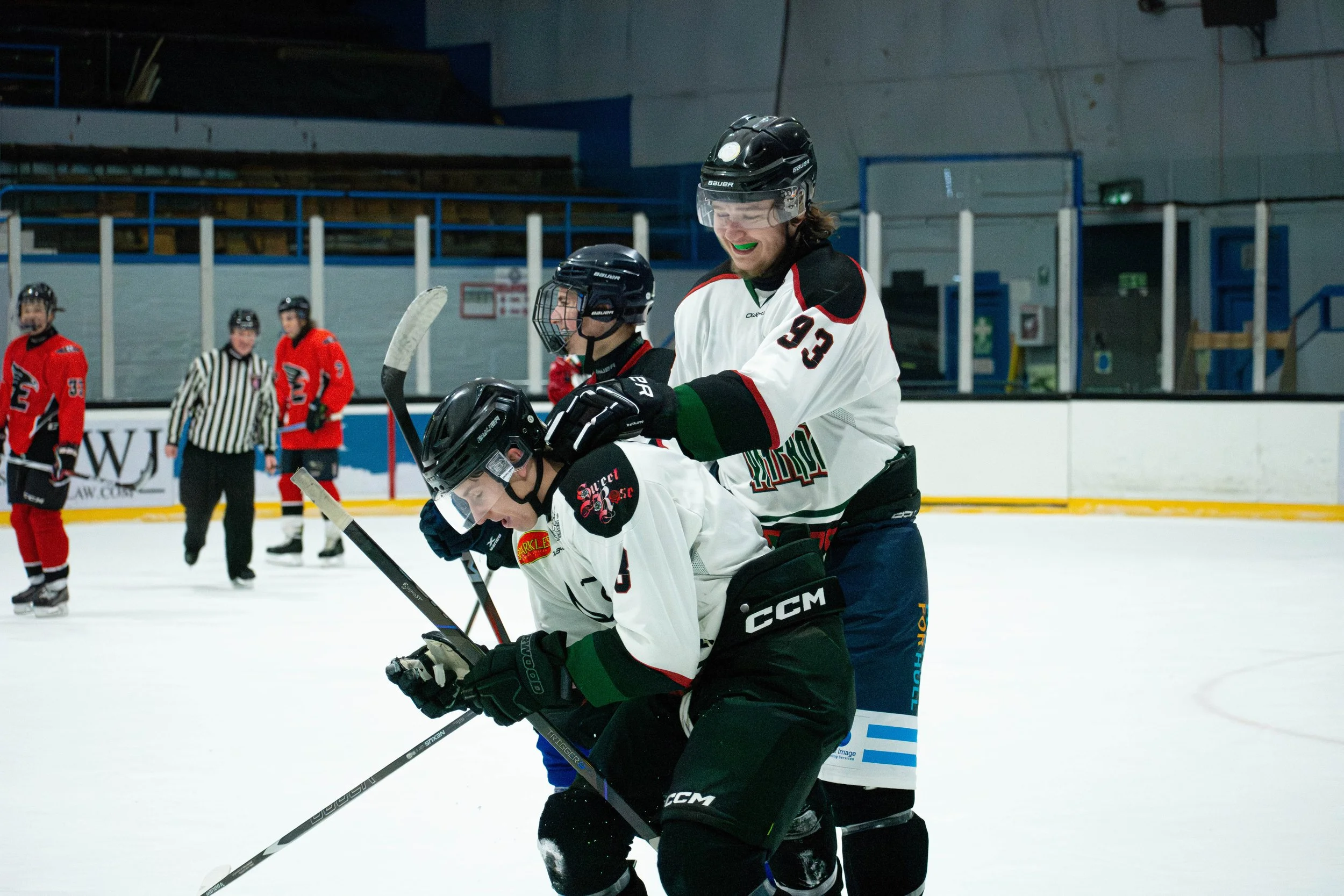 Two ice hockey players in white jerseys celebrate a goal on the ice rink, with other players and a referee in the background.