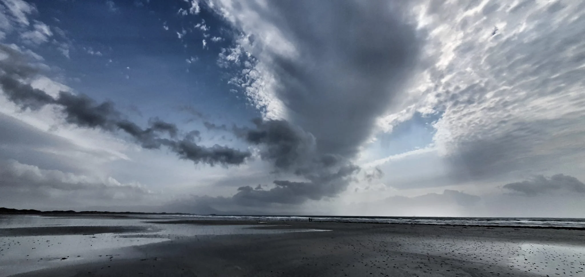 Dark clouds over a sandy beach with water reflecting the sky, and two small people walking in the distance under a partly cloudy sky.