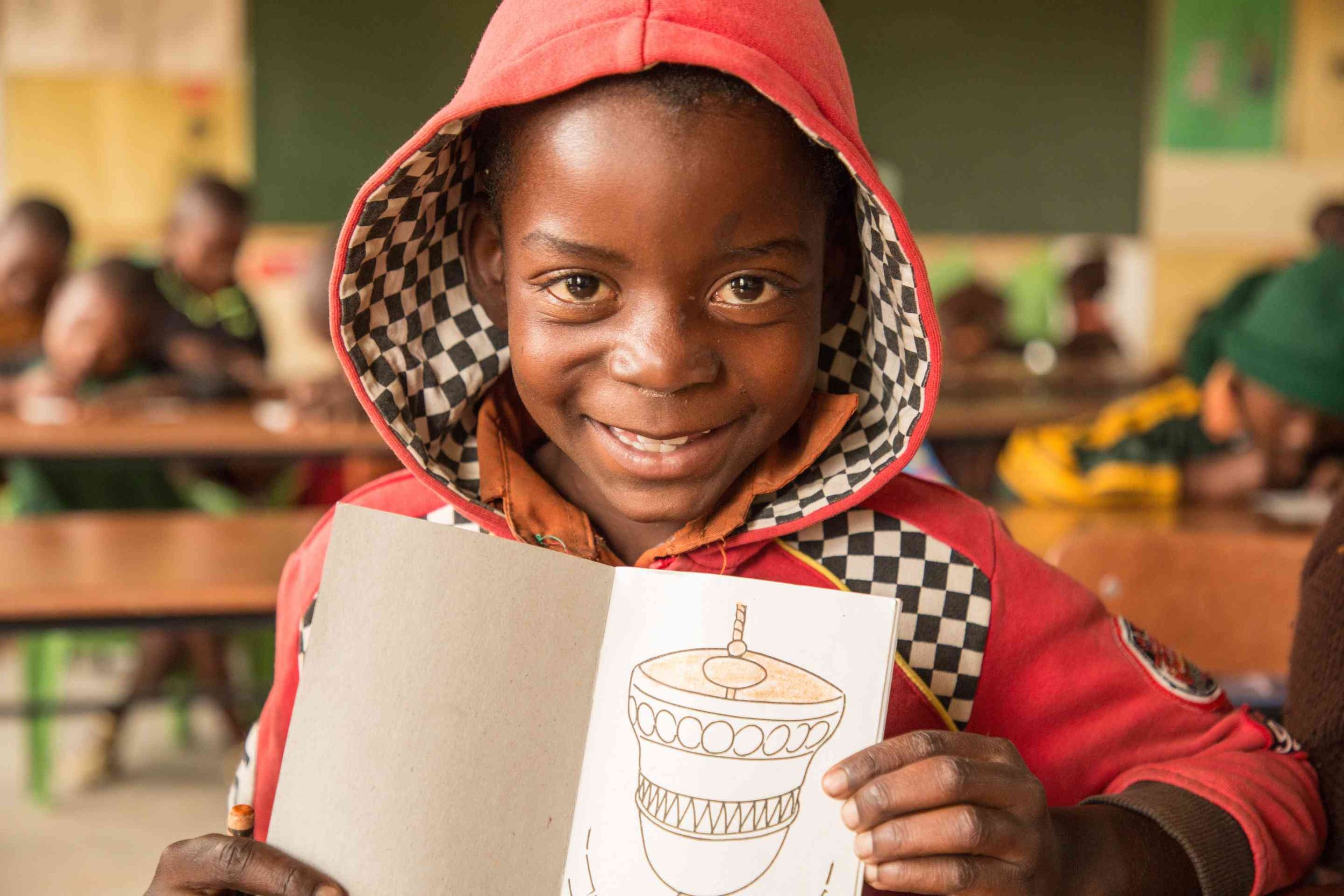 A young boy with a big smile, wearing a red hoodie with a checkered lining, holding an open colouring book with a drawing of a decorated jar or pot, standing in a classroom with other children in the background.