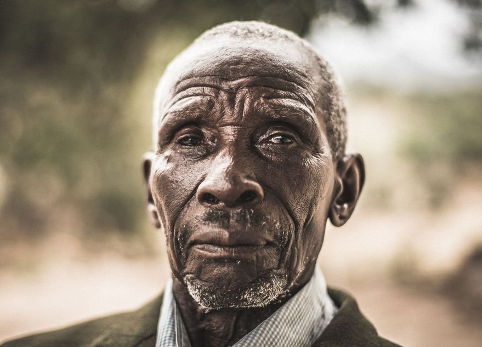 Close-up of an elderly man with dark skin, short gray hair, and a serious expression, wearing a collared shirt and jacket, with a blurred outdoor background.