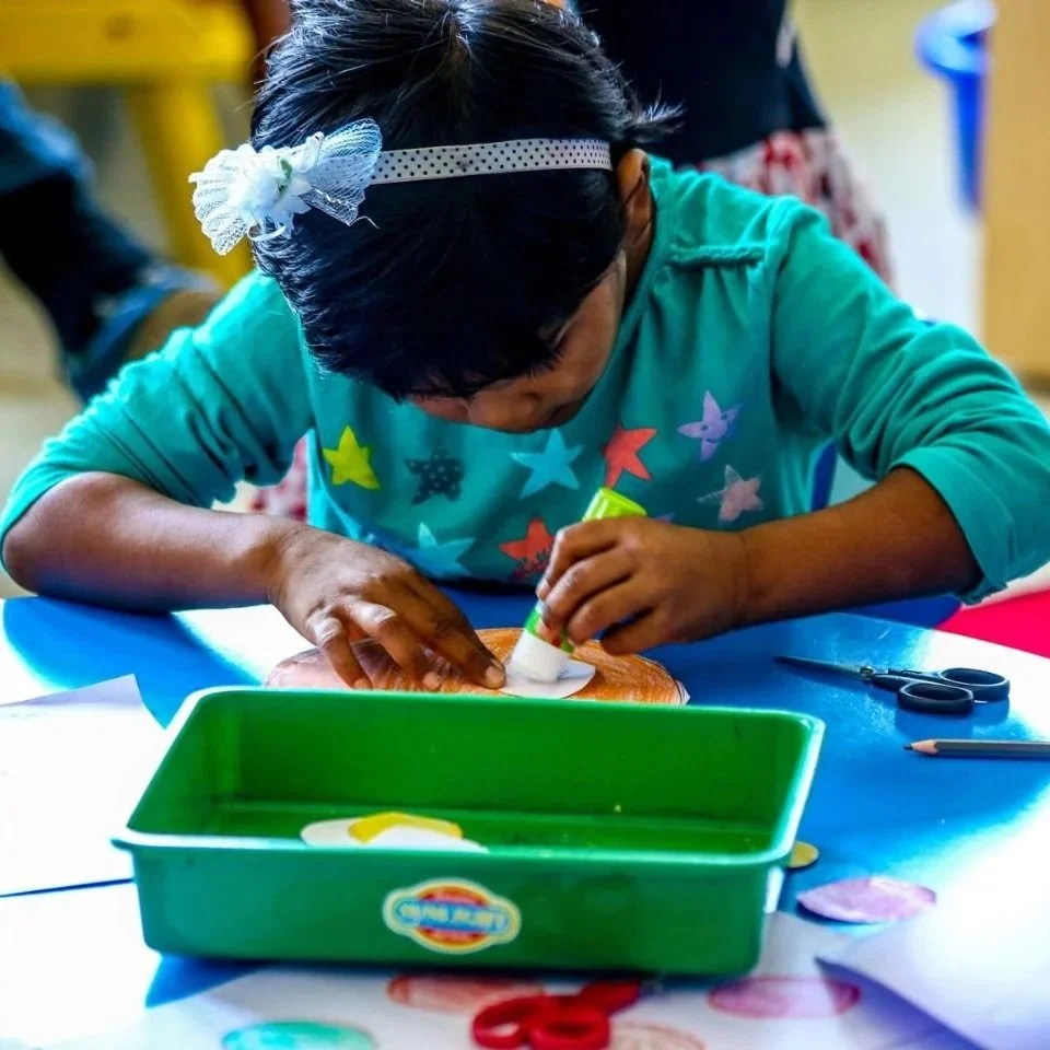 Child girl with a headband and star-patterned shirt engaged in arts and crafts, drawing or gluing on a piece of paper at a table.