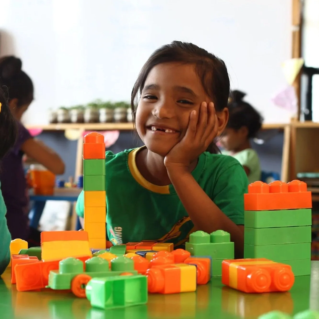 A young girl with a missing front tooth smiling while resting her head on her hand. She is surrounded by colorful building blocks on a table in a classroom or playroom.
