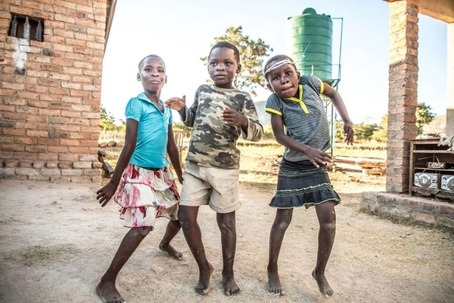 Three children playing and dancing barefoot outside near a brick structure, with a green water tank in the background.