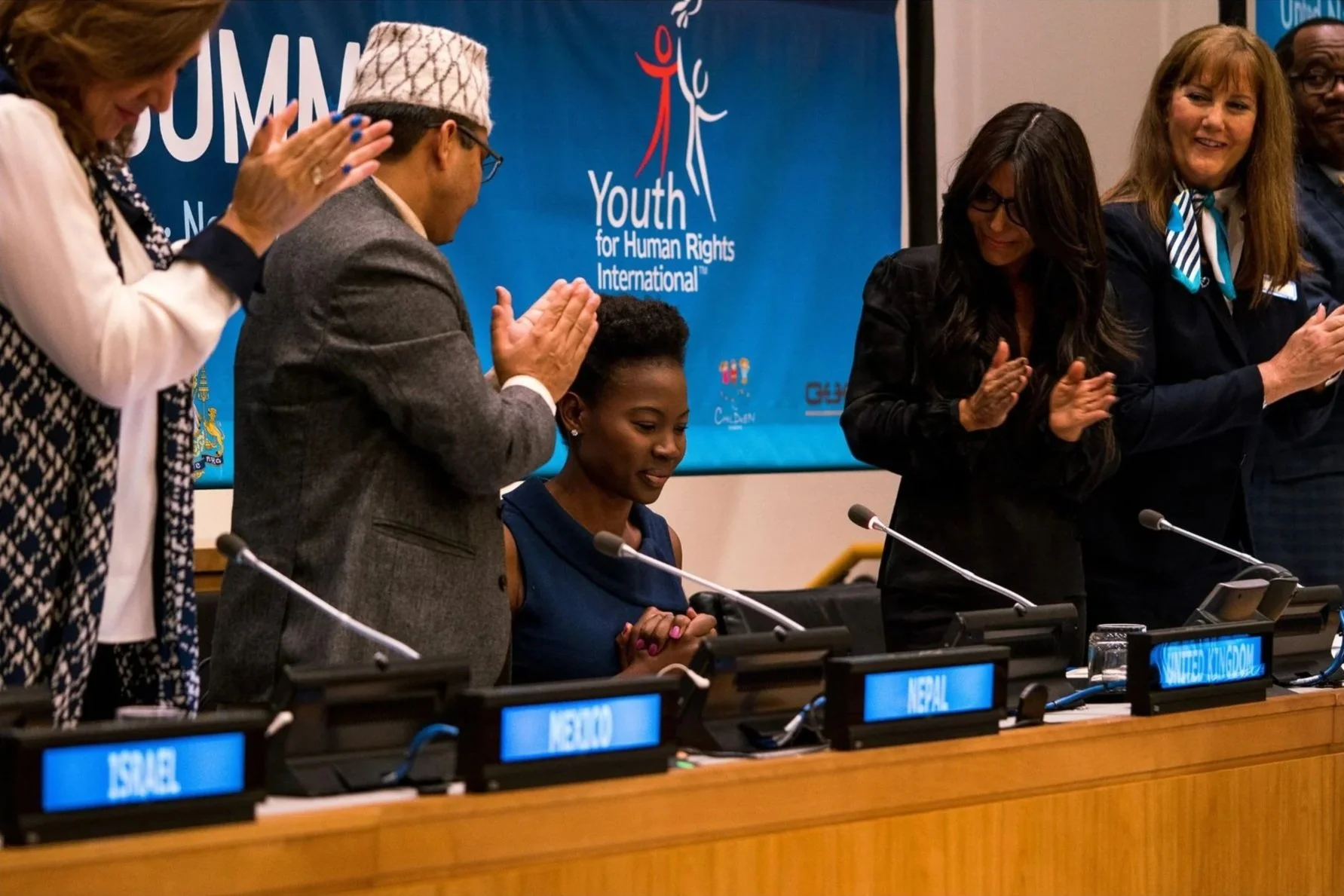 Group of diverse young people and adults standing behind a table, clapping, at a Youth for Human Rights International event, with a blue banner in the background displaying the organization's logo and name, and country nameplates on the table.