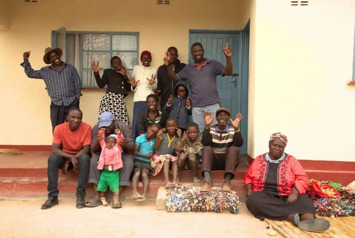 Group of people, including children and adults, posing outside a building with a blue door and window. Some are sitting, some are standing, and they are smiling and making hand gestures. There are colorful items on the ground in front of them.