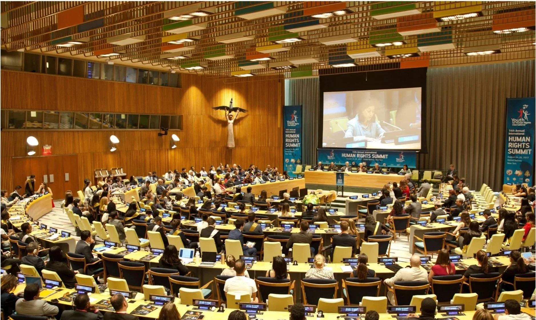 A large conference room during the 14th Annual International Human Rights Summit, with many attendees seated at curved desks, a stage with panelists, and a large screen displaying a speaker.