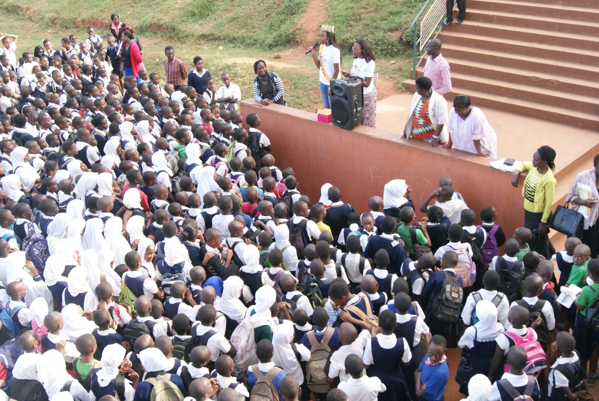 A large group of school children standing outdoors facing a small stage with adults, some of whom are speaking into a microphone. The children wear school uniforms, and the scene appears to be a school assembly or event.