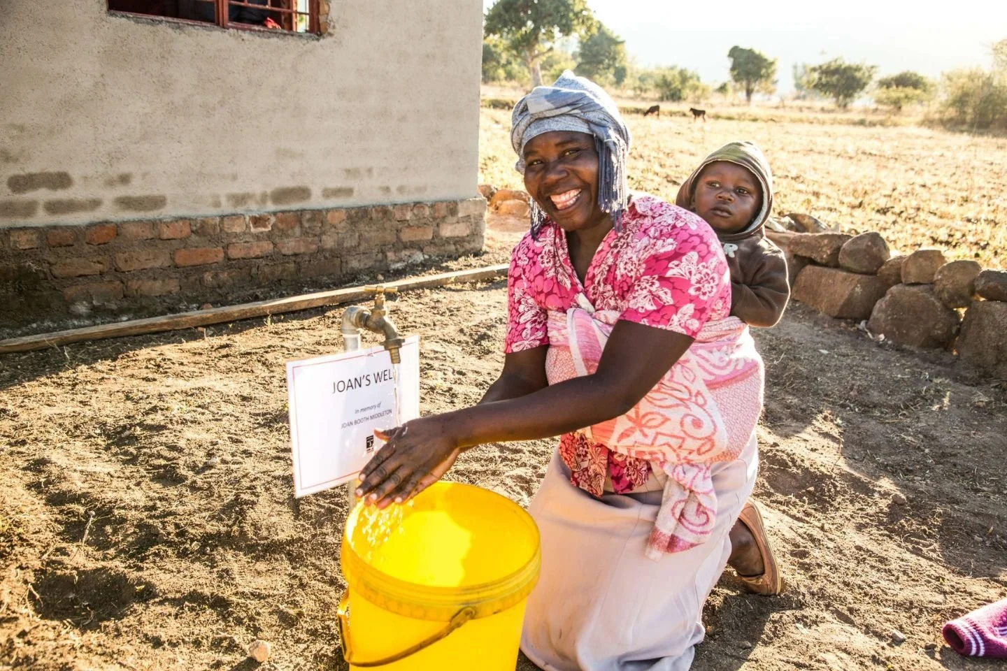 A woman with a child on her back using a water tap connected to a yellow bucket outside a house, smiling at the camera. The background shows an open field with a few trees and animals.