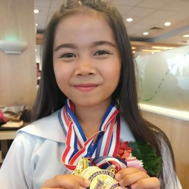 Young girl with long dark hair smiling, wearing a white shirt and medals around her neck, holding a bouquet of flowers, in an indoor setting.