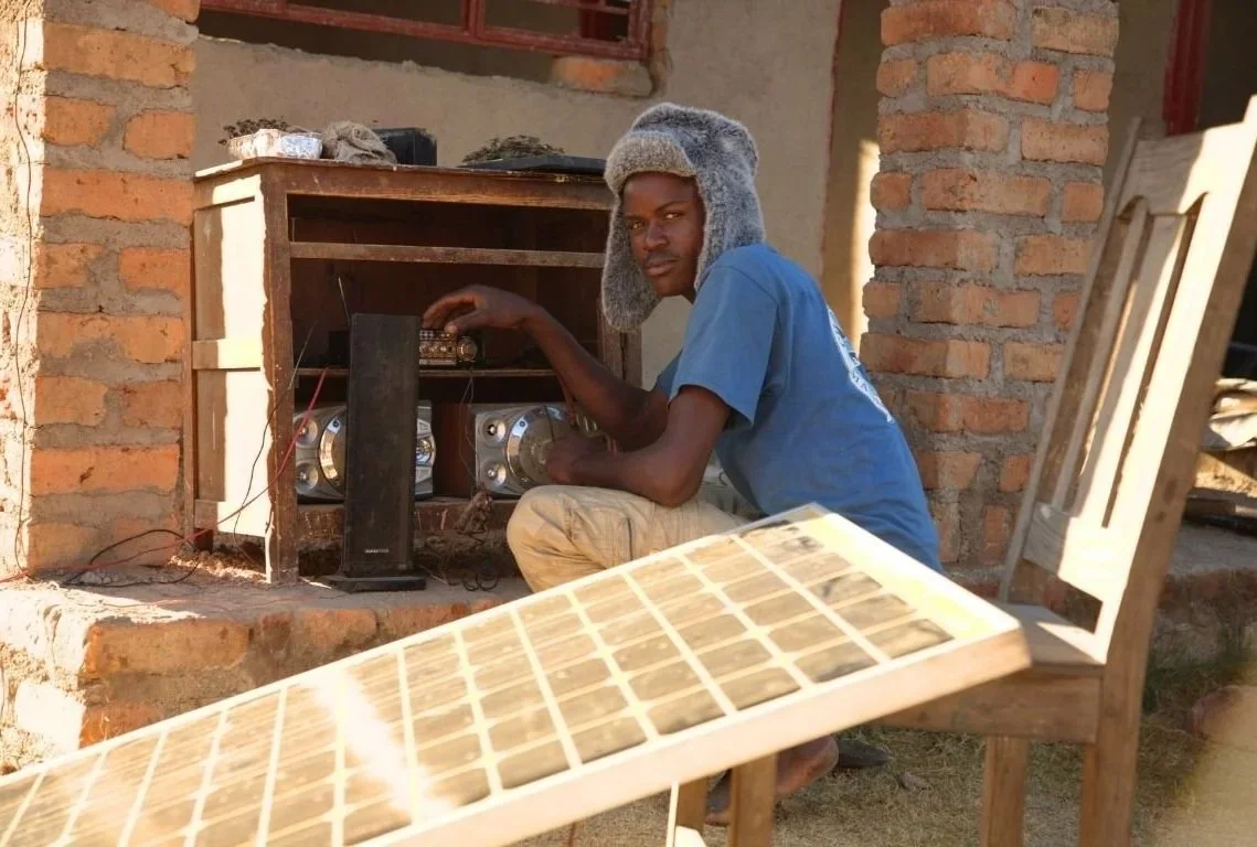 Young man with a warm hat working on a homemade radio inside a brick structure, sunny outdoor setting.