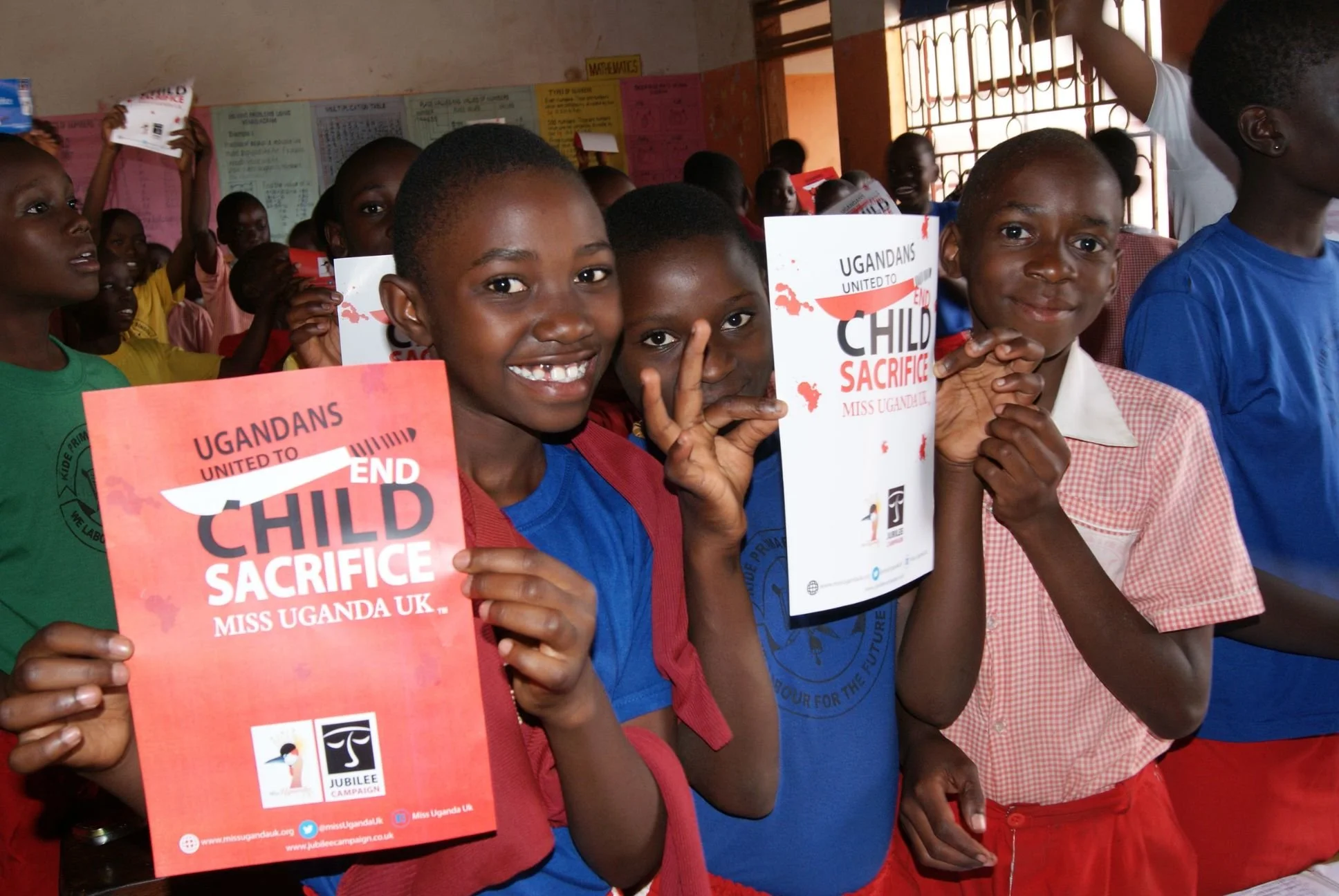 Group of children holding signs that read 'Ugandans United to End Child Sacrifice Miss Uganda UK' during a rally or event.